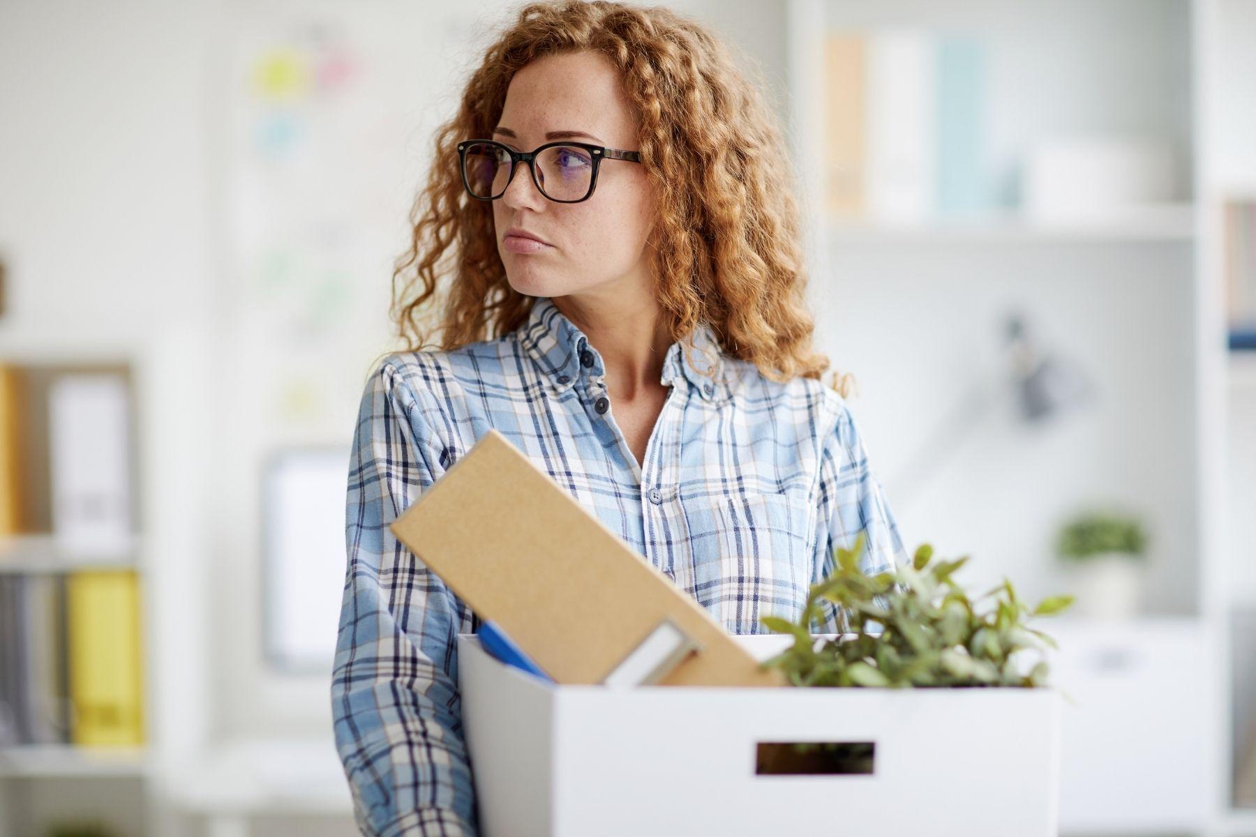 Photo of a woman in an office. She is holding a box with her personal items in it.