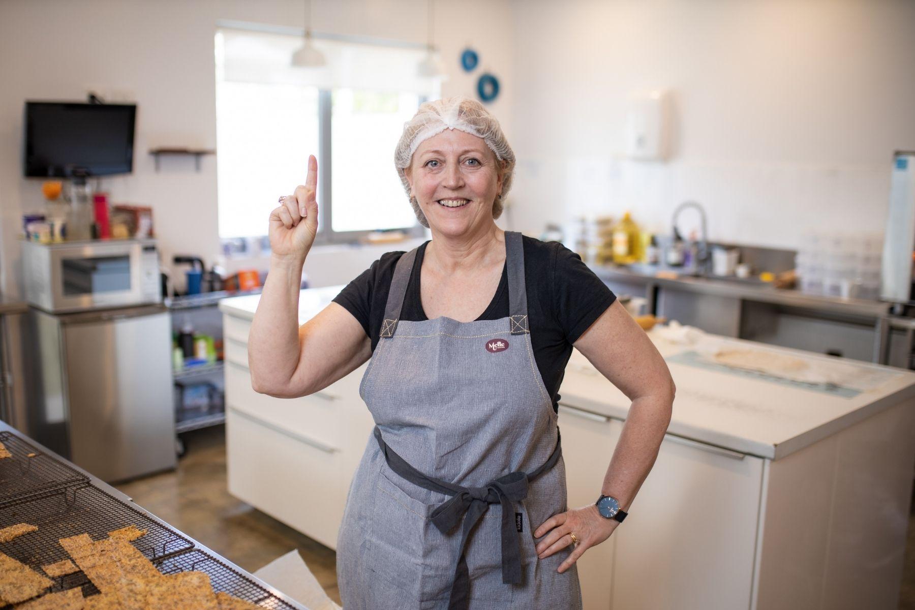 Image of a woman in a kitchen with an apron and a hair net looking at the camera with her hand on her hip and a finger raised in the air.