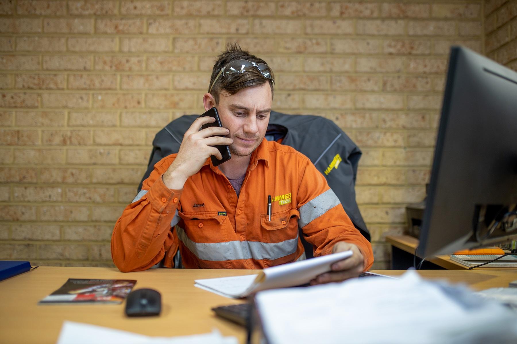 Photo of a male business owner. He is sitting at a desk, reviewing documentation while he is on the telephone.