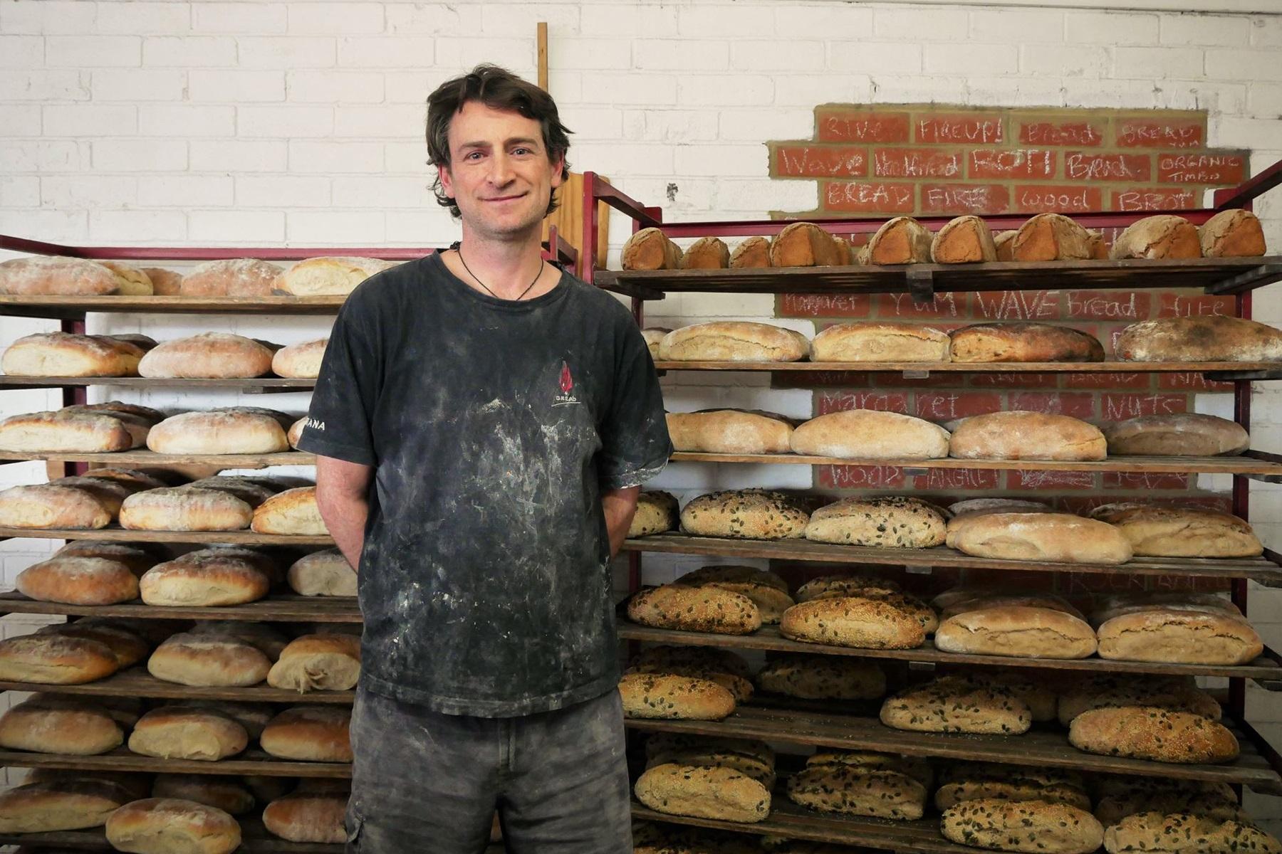 Photo of Santana from Margaret River Woodfired Bread. He is standing in front of shelves filled with bread.