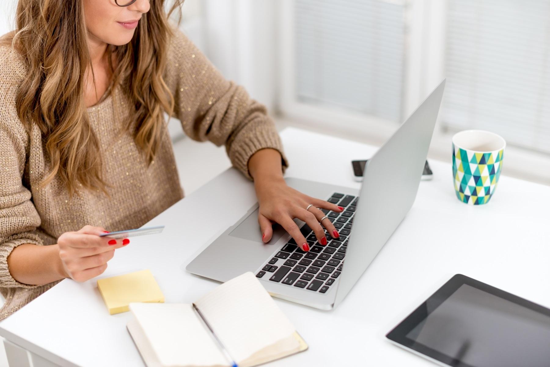 Photo of a woman sitting at a desk while using a laptop. She is holding a credit card while she completes an online payment.