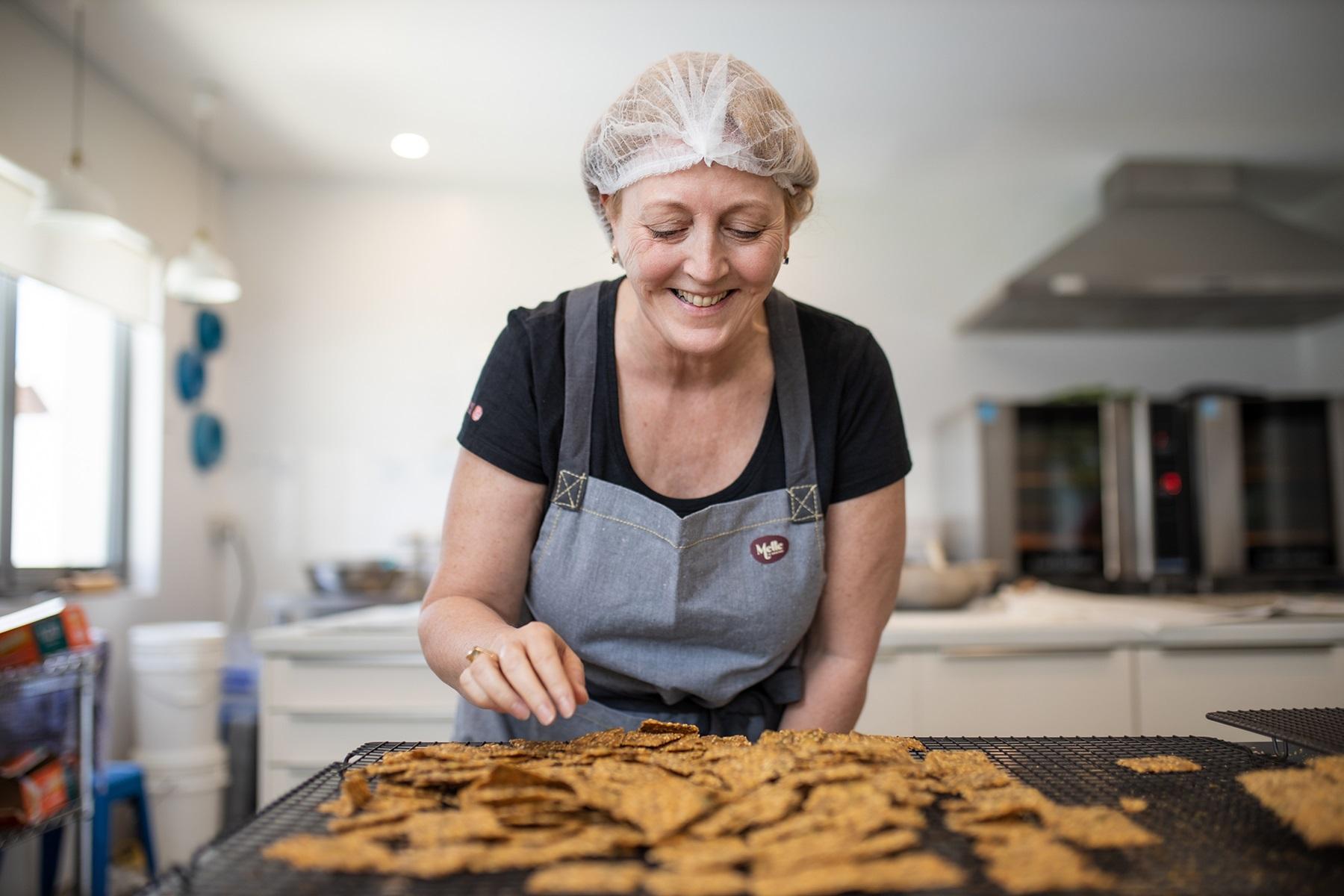 Photo of  a woman in a commercial kitchen. She is inspecting a baking rack full of lavosh crackers that she has baked.