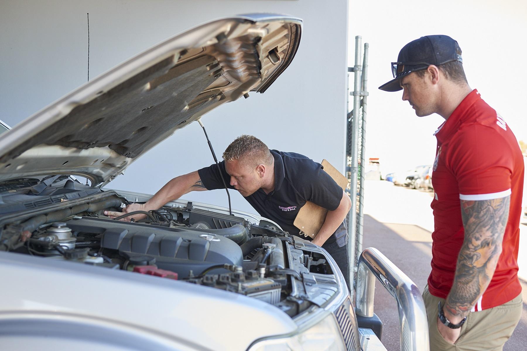 Photo of a male car mechanic looking under the bonnet of a car as a male customer looks on.