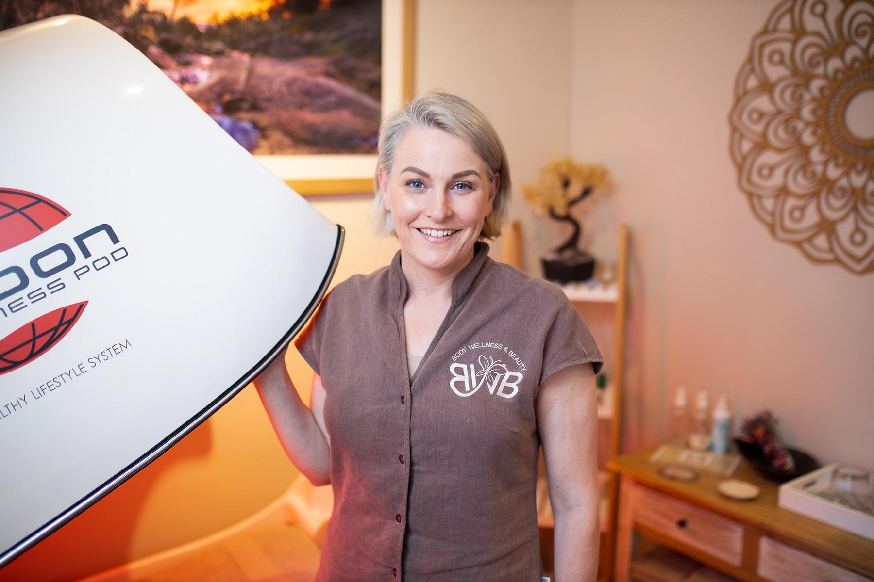 Photo of a female owner of a body wellness and beauty business. She is standing next to a fitness pod machine.