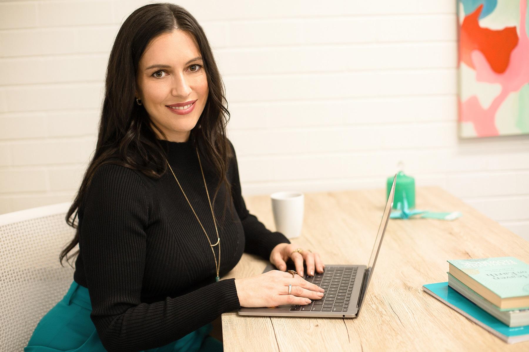 Photo of Kasia McNaught. She is sitting at a desk with a laptop and smiling at the camera.