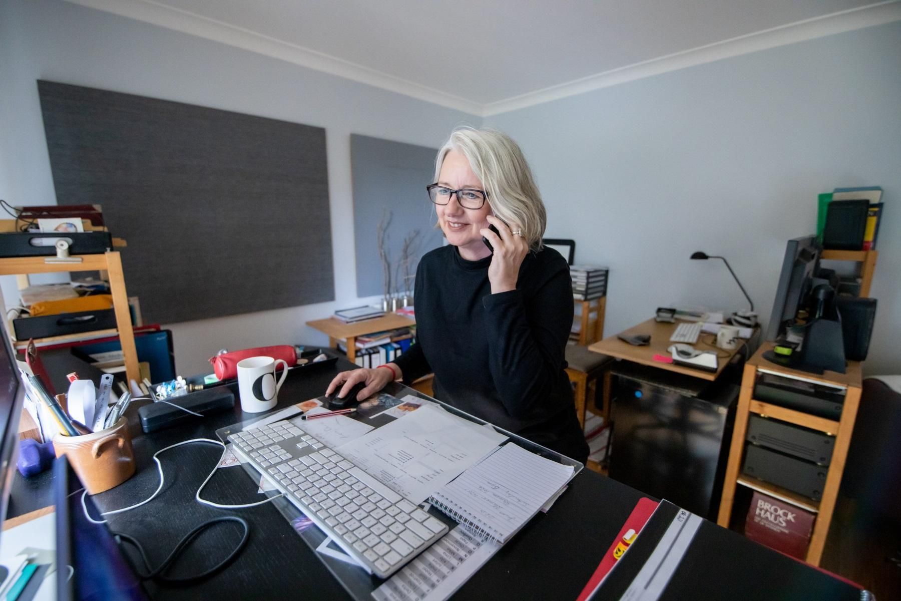 Photo of a woman working in a home office. She is sitting in front of a computer and is talking on her mobile phone.