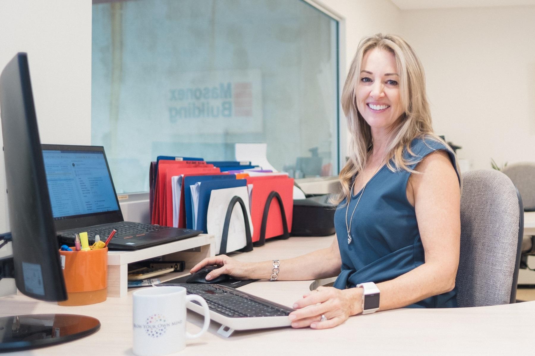 Photo of a woman sitting at a desk with a computer and files around her. She is smiling at the camera.