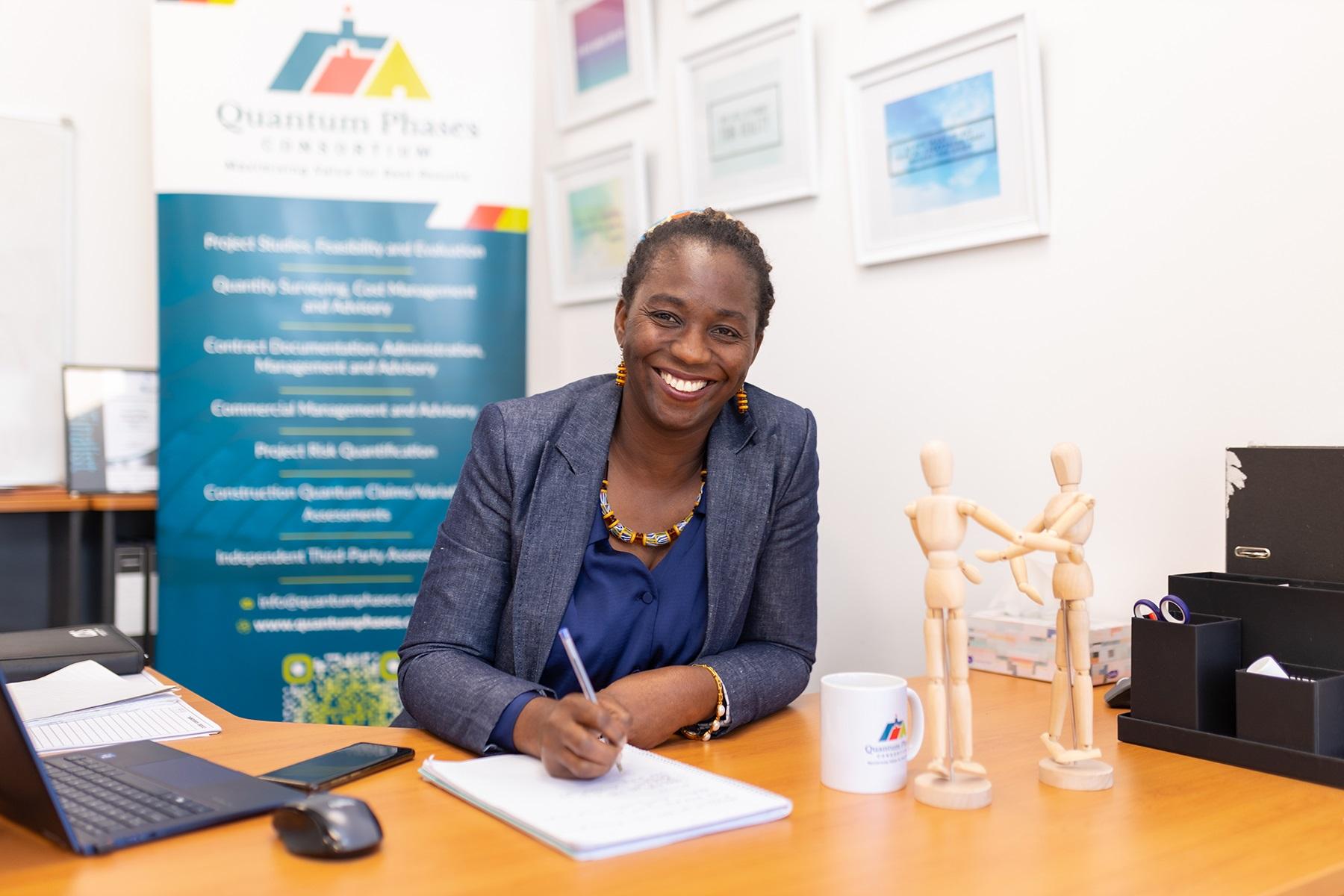 Photo of Adwoa Abban, owner of Quantum Phases Consortium. Adwoa is sitting at a desk with a laptop, notebook and pen and is smiling at the camera.