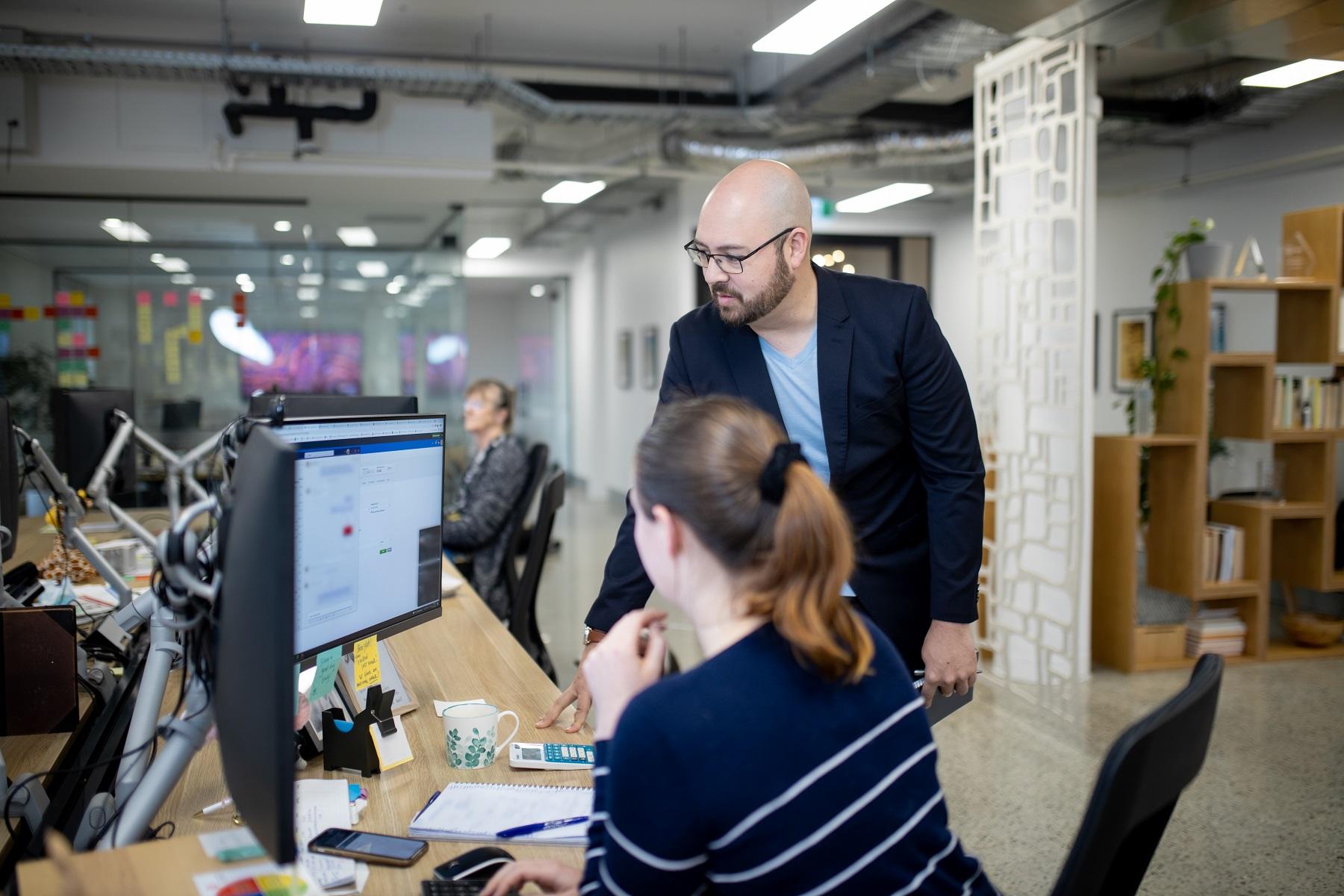 Photo of a man reading from a computer monitor. He is standing next to a female coworker who is sitting at a computer  and reading from the same monitor.