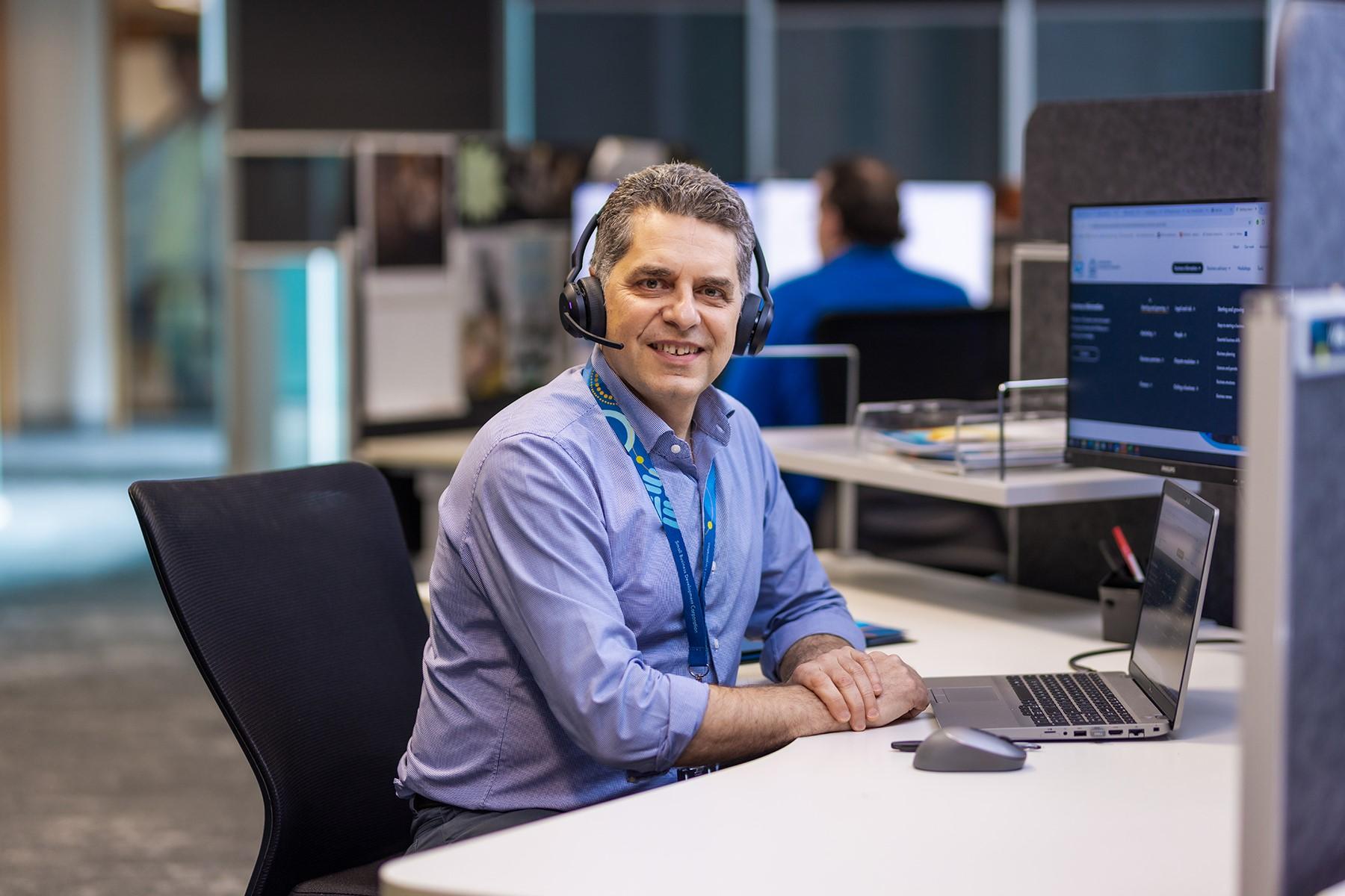 Photo of a male SBDC staff member. He is sitting at a desk, in front of a computer and is wearing a telephone head set.