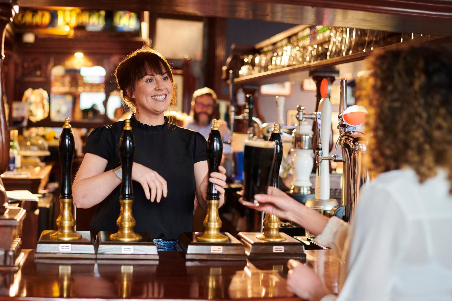 Photo of a woman working in a pub. She is behind a bar, handing a drink over to a customer.