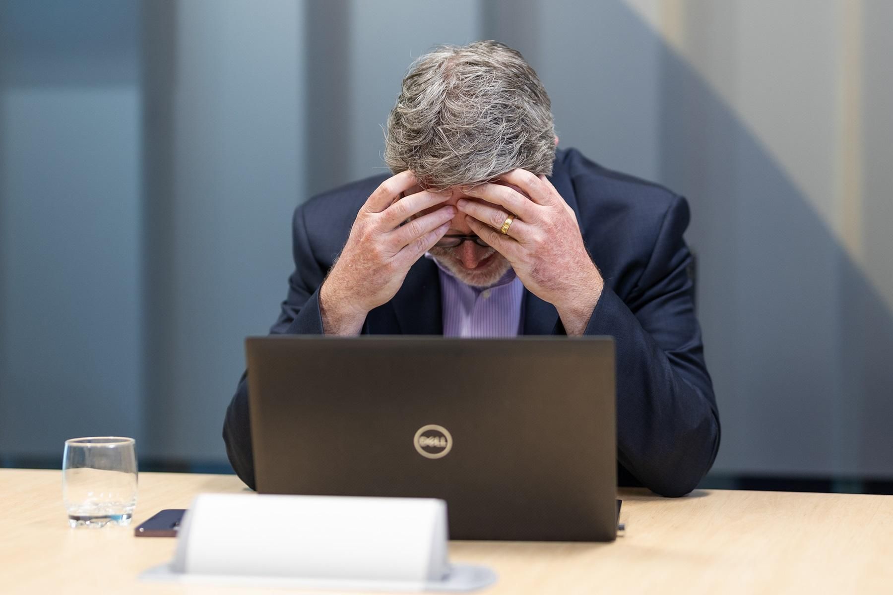 A photo of a man sitting in front of a laptop. He has his head in his hands.