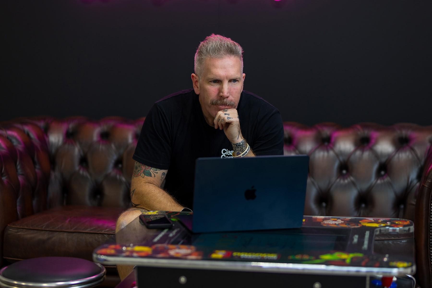 A man sitting on a couch. He is reading from a laptop.