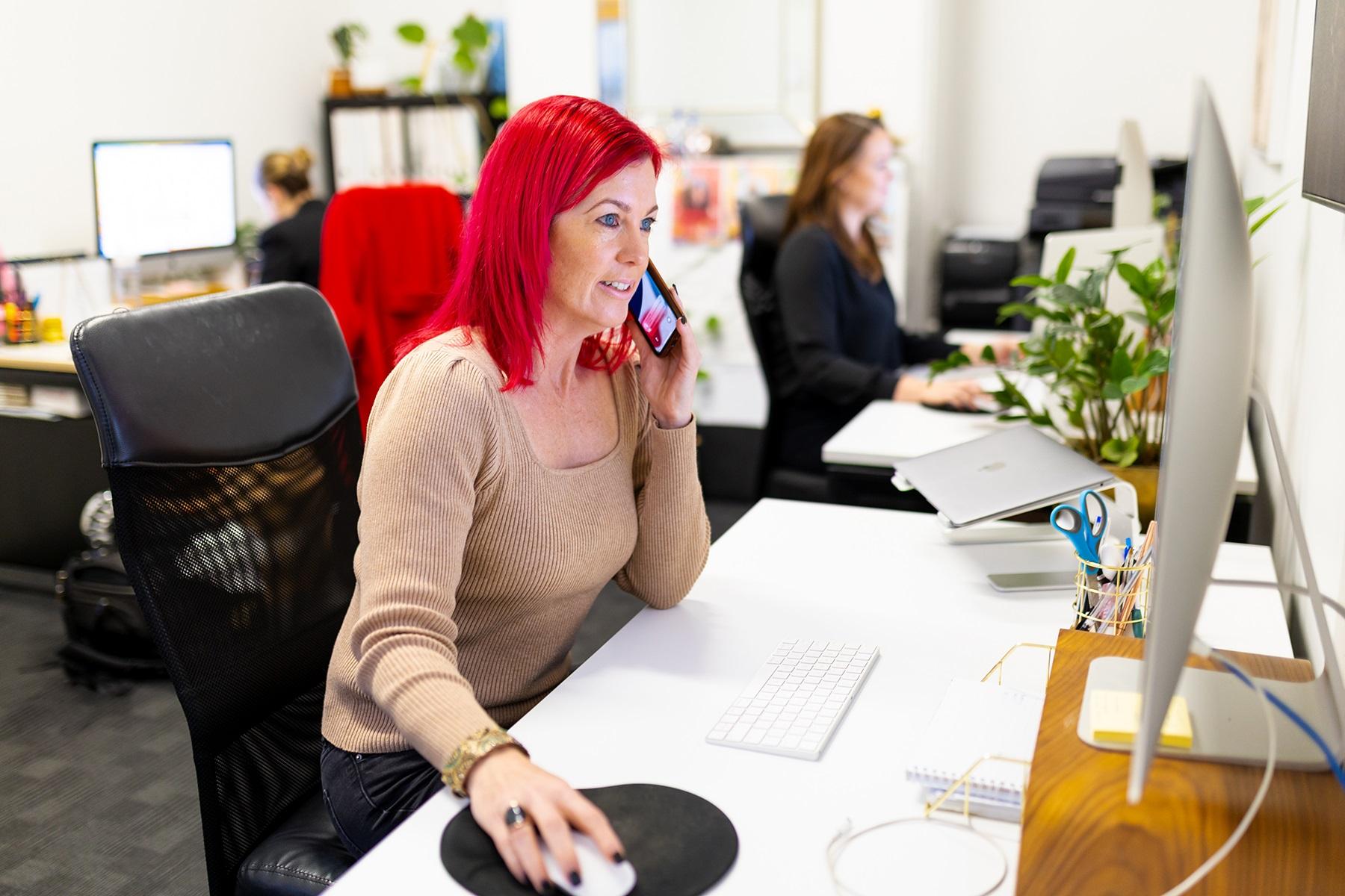 Image of a woman sitting at a desk with a computer using her mobile phone.
