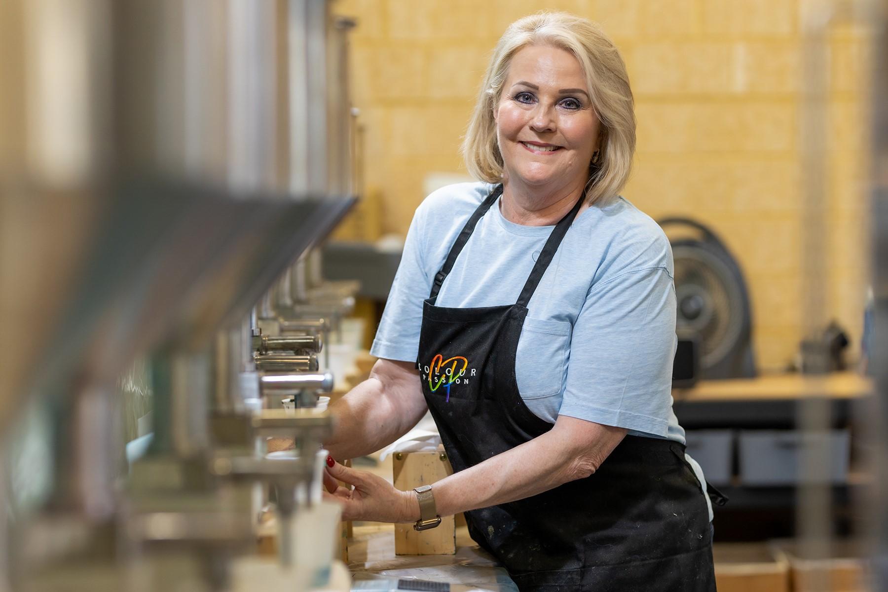 Photo of a women in her business premises. She is standing in front of a row of storage vats which hold high quality pigments and resin supplies.