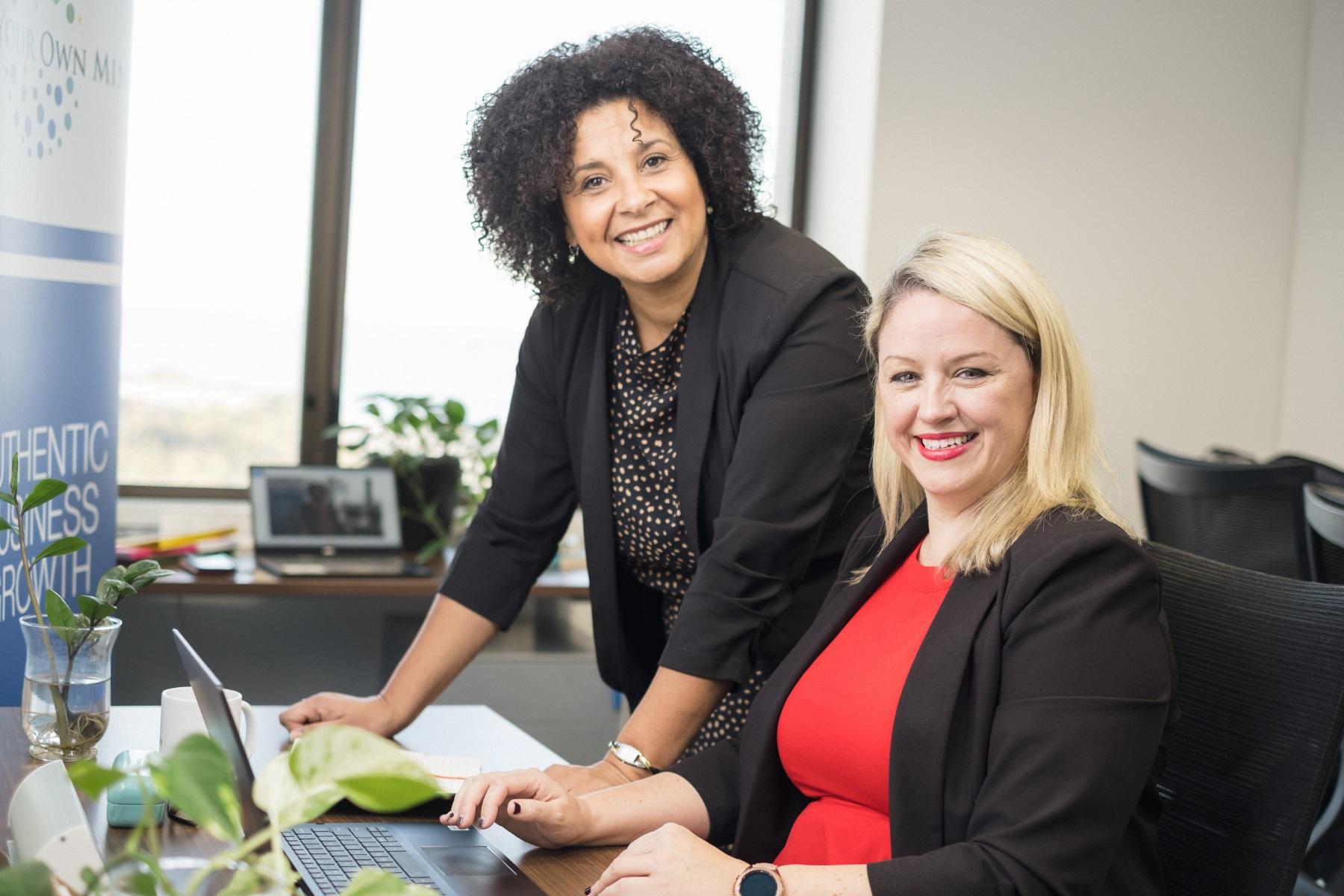 Photo of two female business partners. One is sitting at a desk and the other is standing. They are both smiling at the camera.