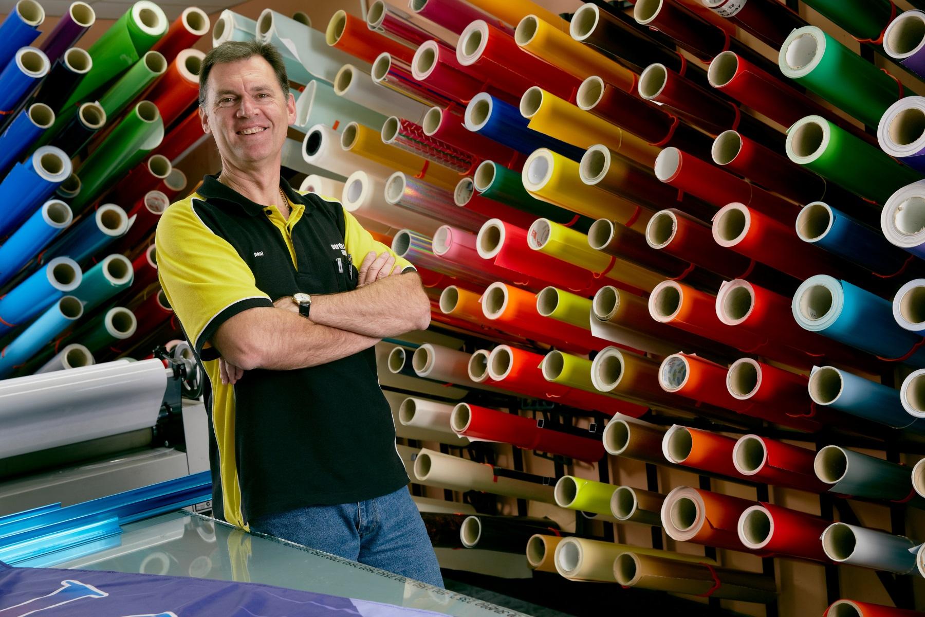 Photo of a man standing in a printing business with rolls of vinyl behind him.