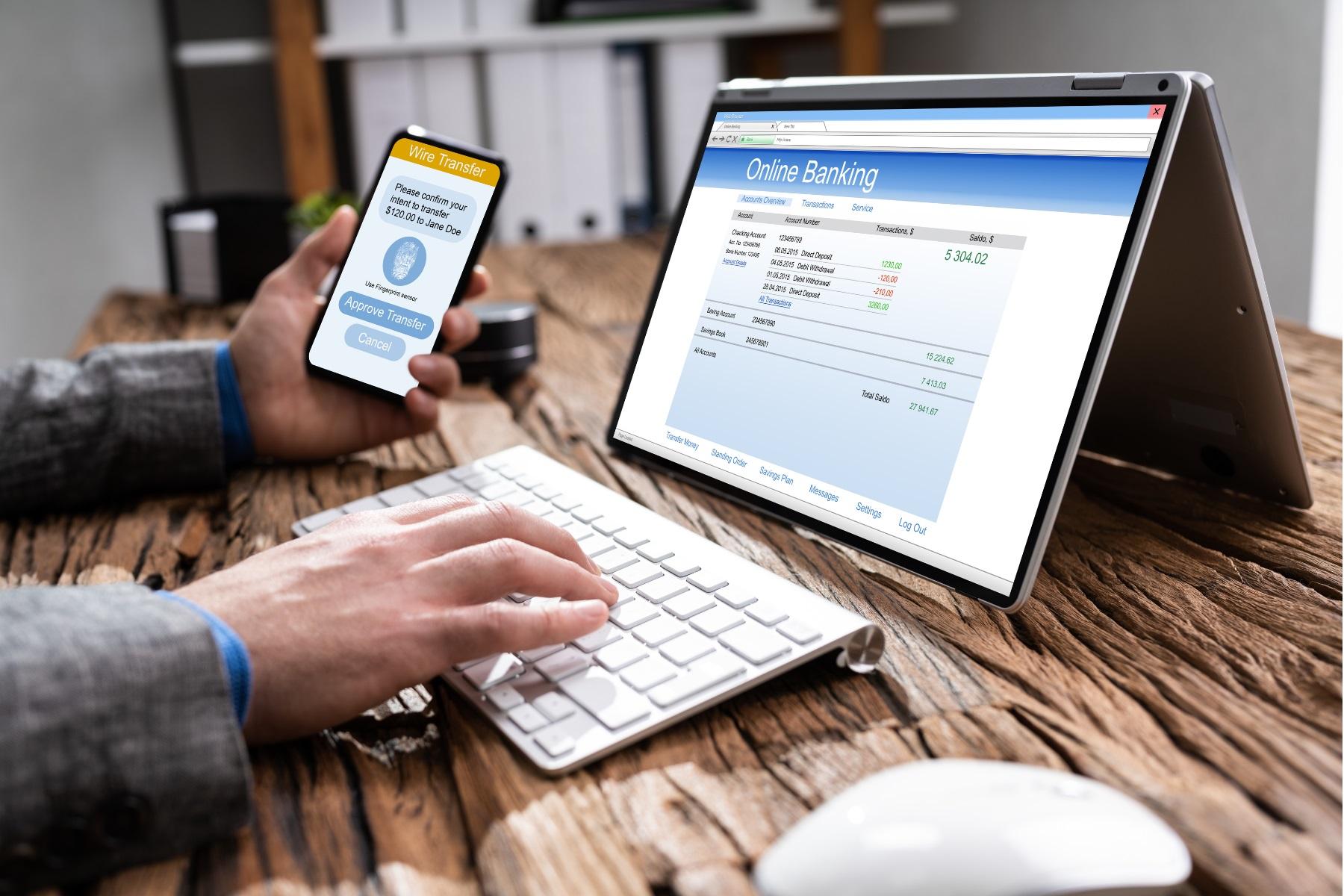 Photo of a person sitting at a table. They are using a mobile phone to complete two factor authentication to authorise an online banking payment being made through a laptop.