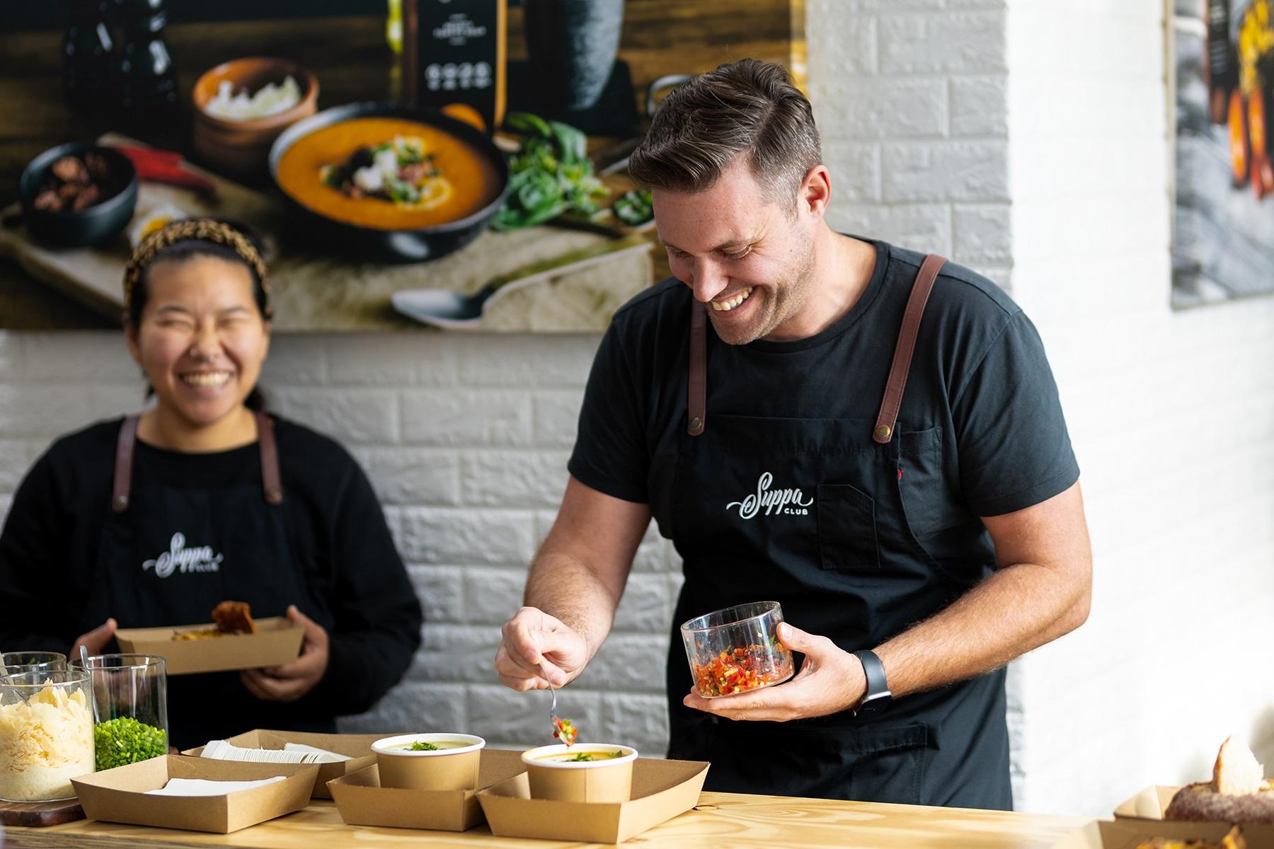 Photo of a male business owner preparing orders of soup for customers in his food based business. A staff member is in the background of the photo holding a container of food.