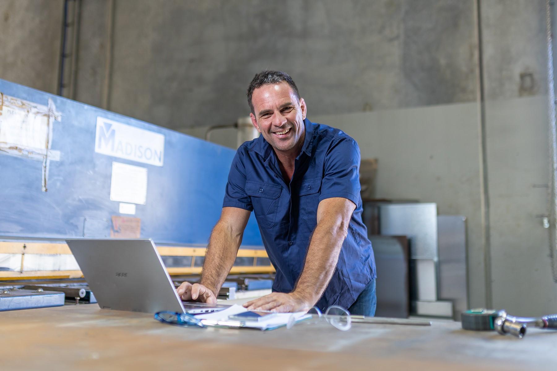 Photo of a male business owner in a fabrication workshop. He is standing at a desk and has a laptop in front of him.