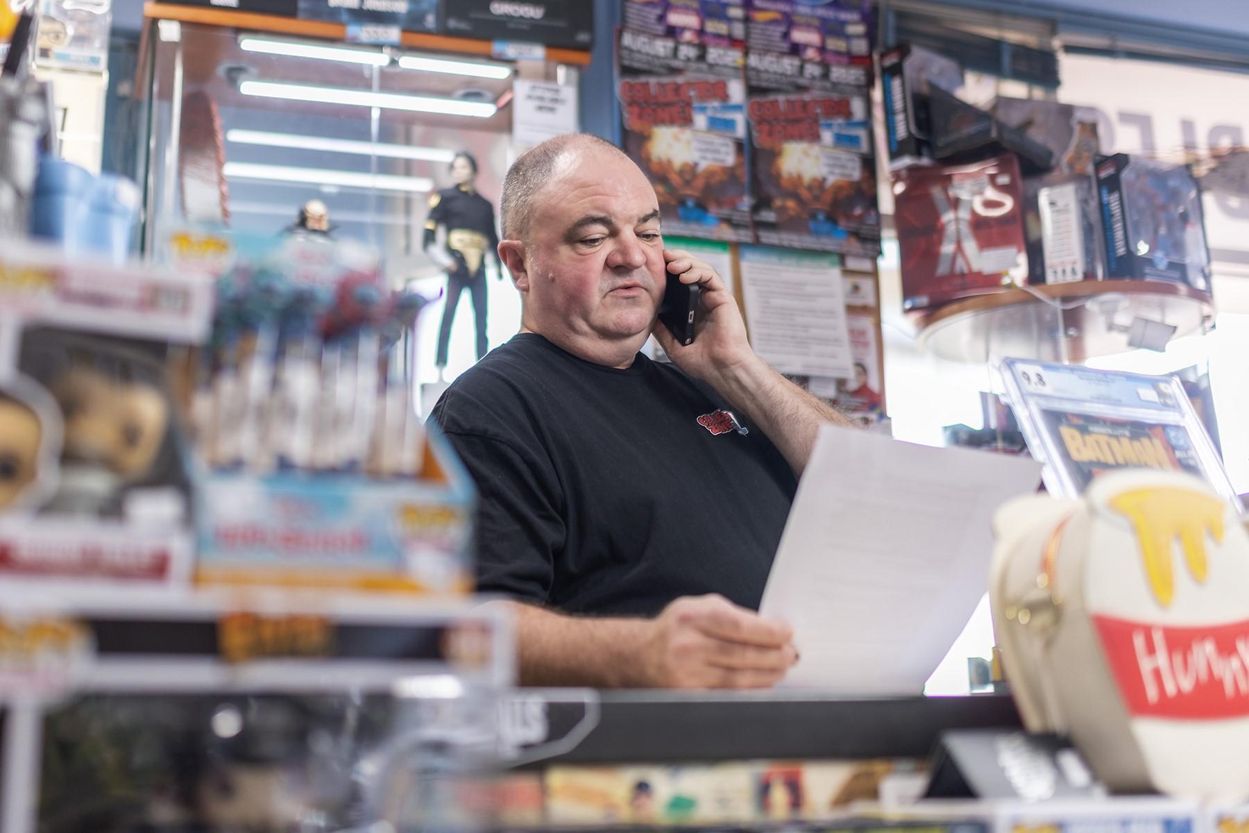 Photo of a male business owner. He is standing behind the counter of his retail store and is reading a piece of paper as he speaks on the phone.