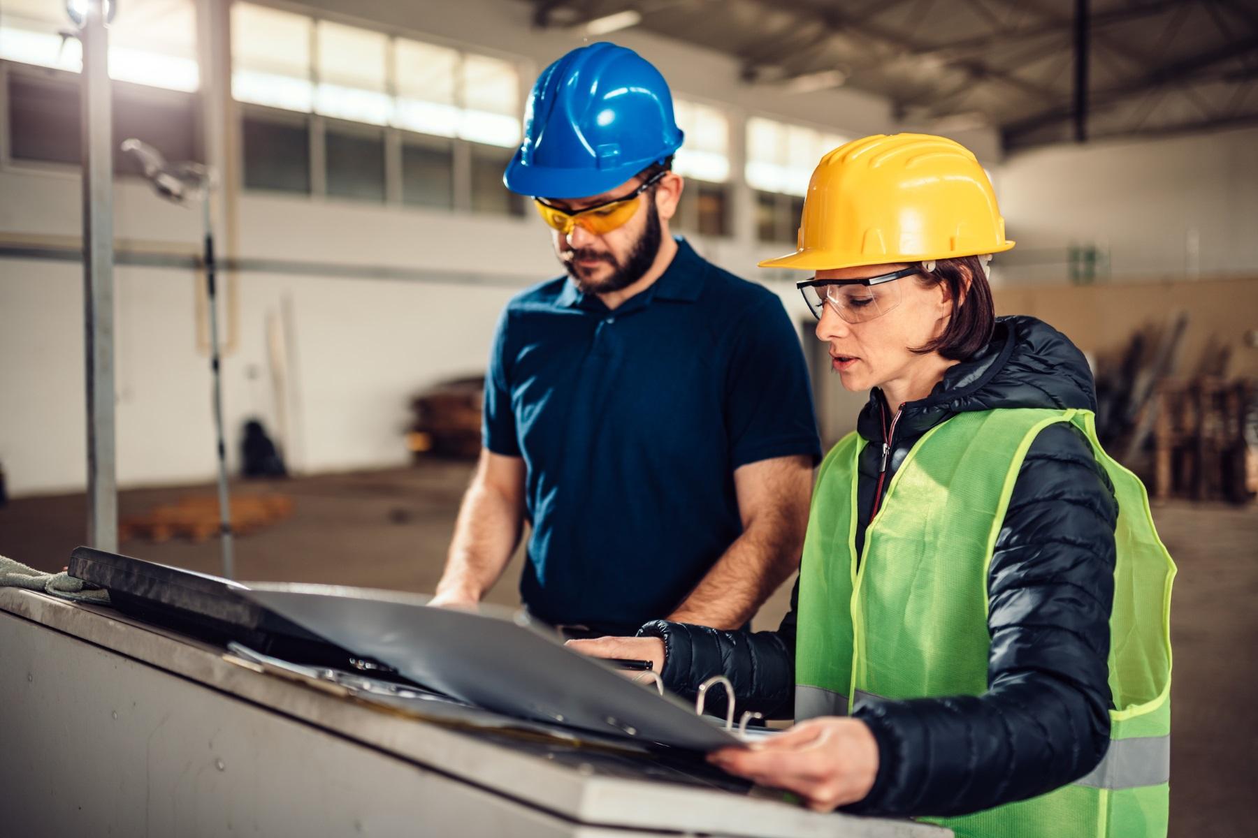 Photo of a woman and man standing in a workshop. They are wearing safety gear and reviewing documentation.