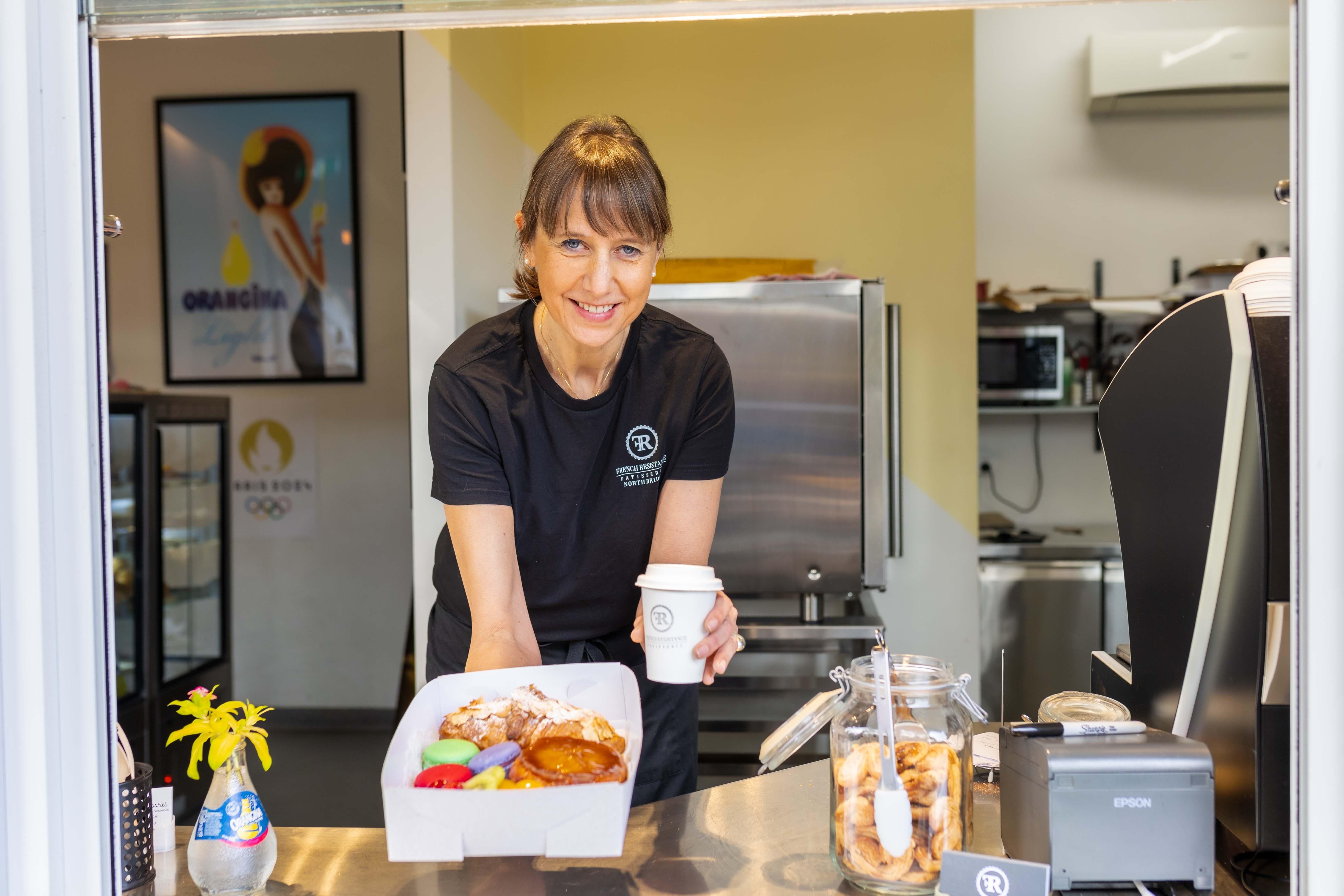 Photo of Gaelle Morival, owner of the French Resistance cafe in Northbridge. She is behind the counter of her cafe, holding a box of pastries and a coffee.