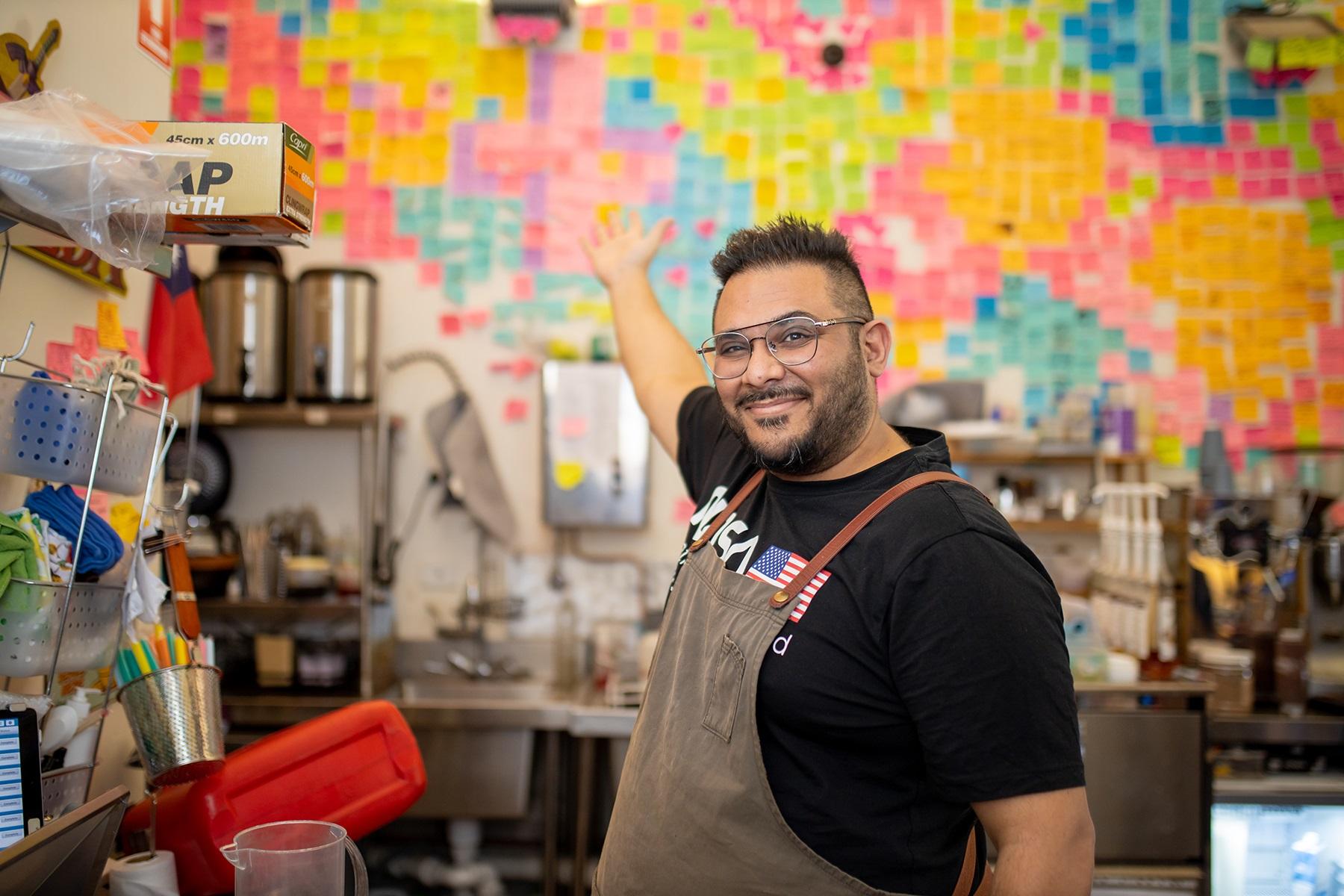 Photo of Dharmesh Patel pointing to the wall of post-it notes on the wall of his cafe, Game City Espresso.