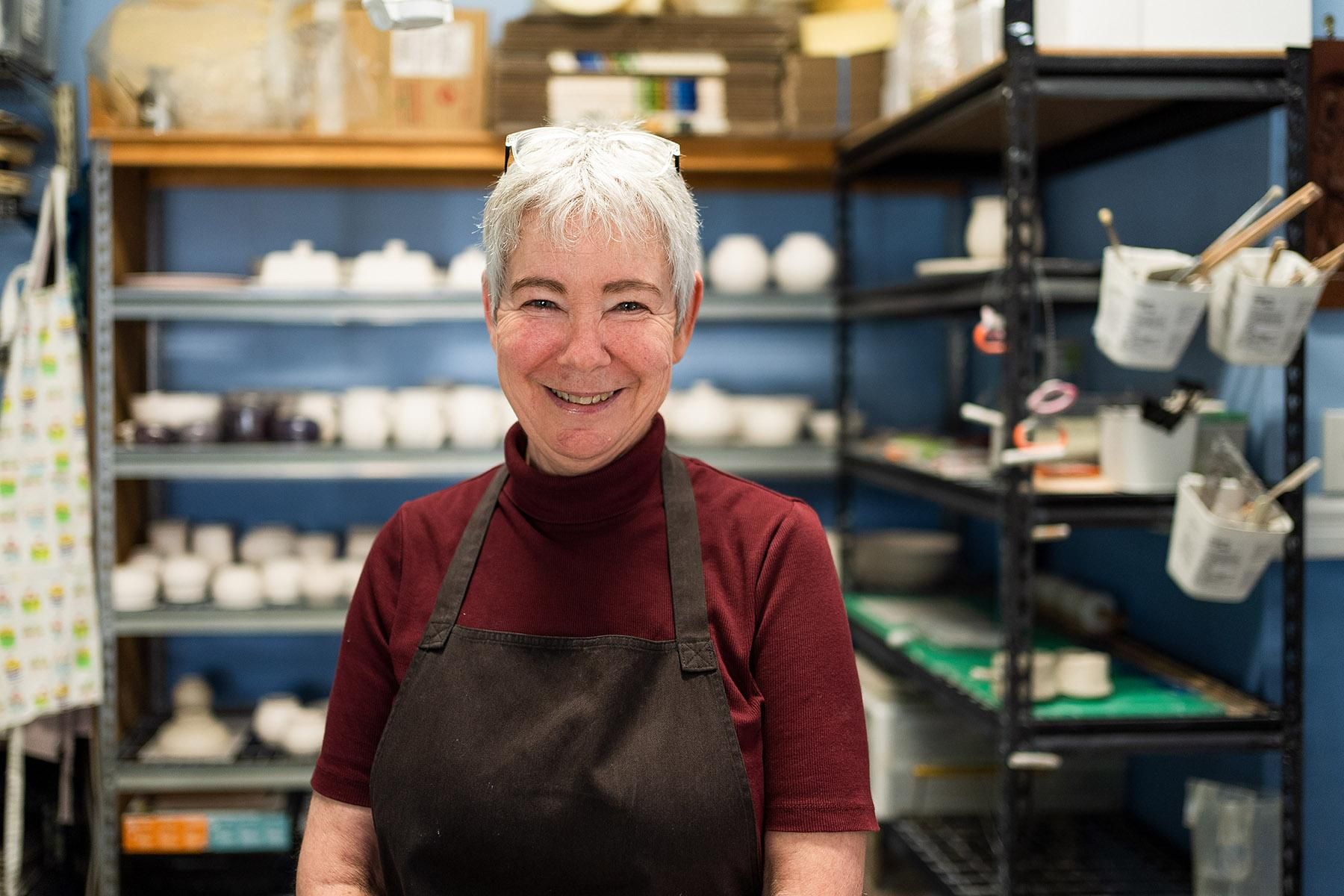Photo of Vicki Read, owner of Torbay Pottery. She is standing in front of shelves which have pottery items on them.