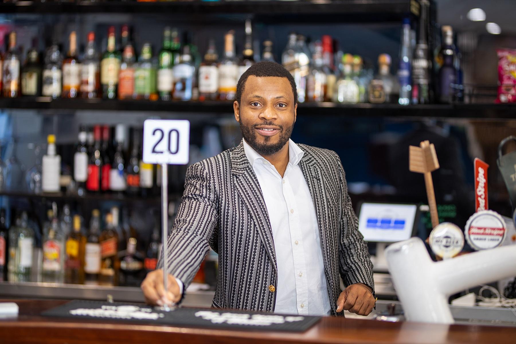 Photo of a male bar and restaurant owner. He is standing behind the bar and is holding a table number to give to a customer.