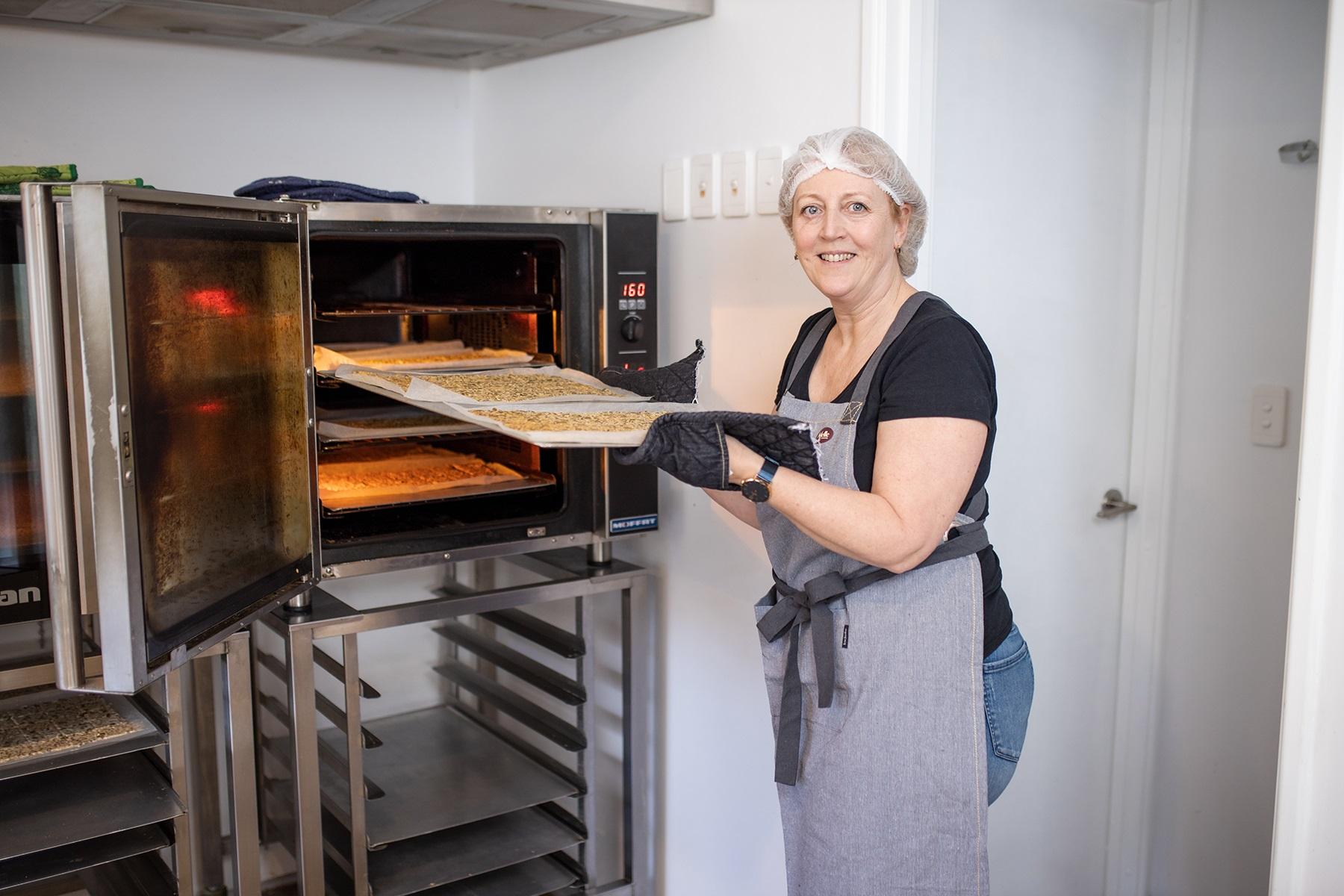 Photo of a woman in a commercial kitchen. She is wearing a hair net and apron and is putting a tray of dough into the oven.