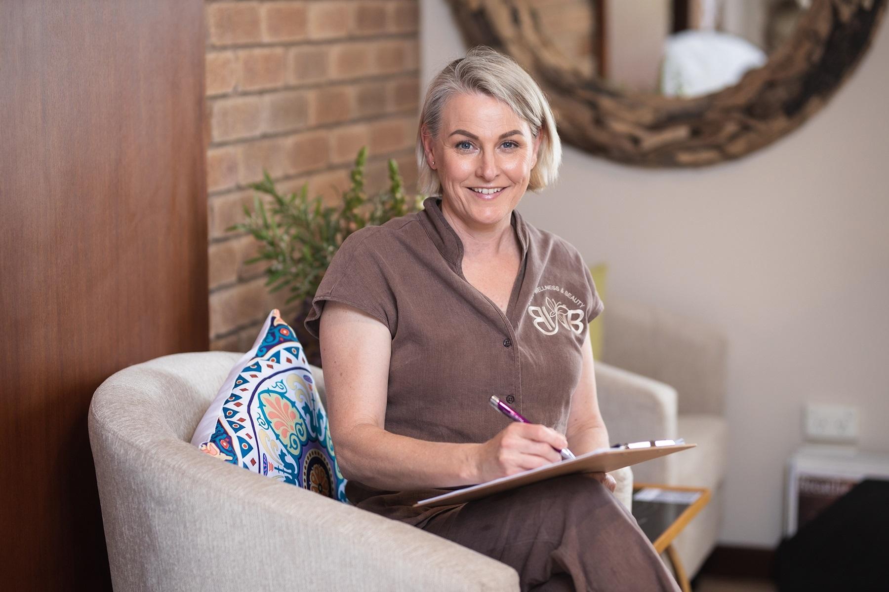 Photo of a female business owner. She is sitting in a tub chair and has a clipboard in hand that she is writing on.