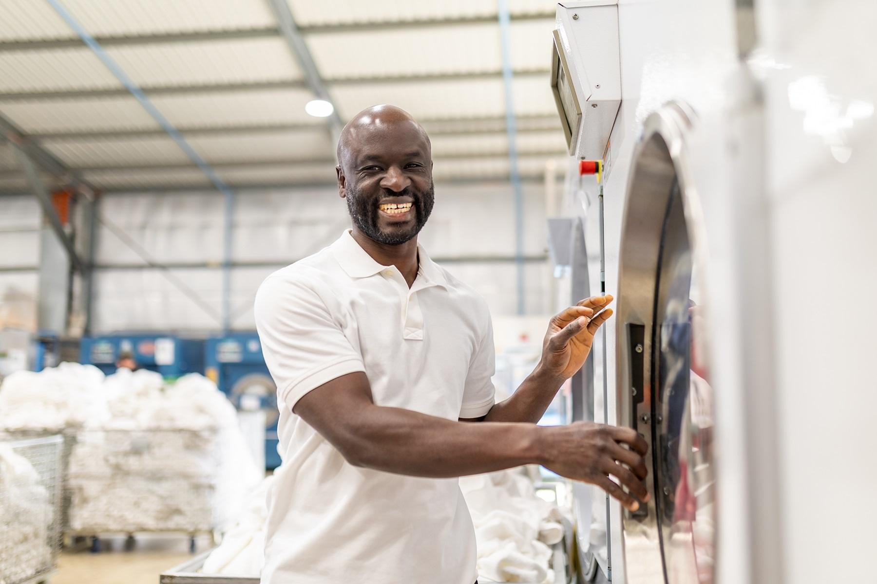 Photo of Ezekial Amudala, who prefers to be known as Mash, in front of a commercial washing machine in his business Baythe Linen and Laundry.
