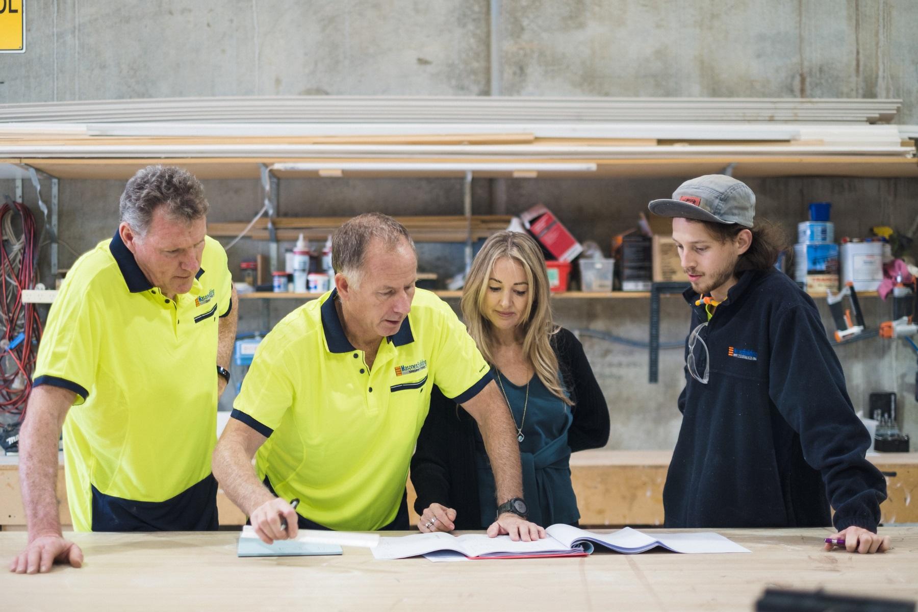A photo of a male and female business partner discussing plans with their male employees at work bench in their industrial fitout workshop.
