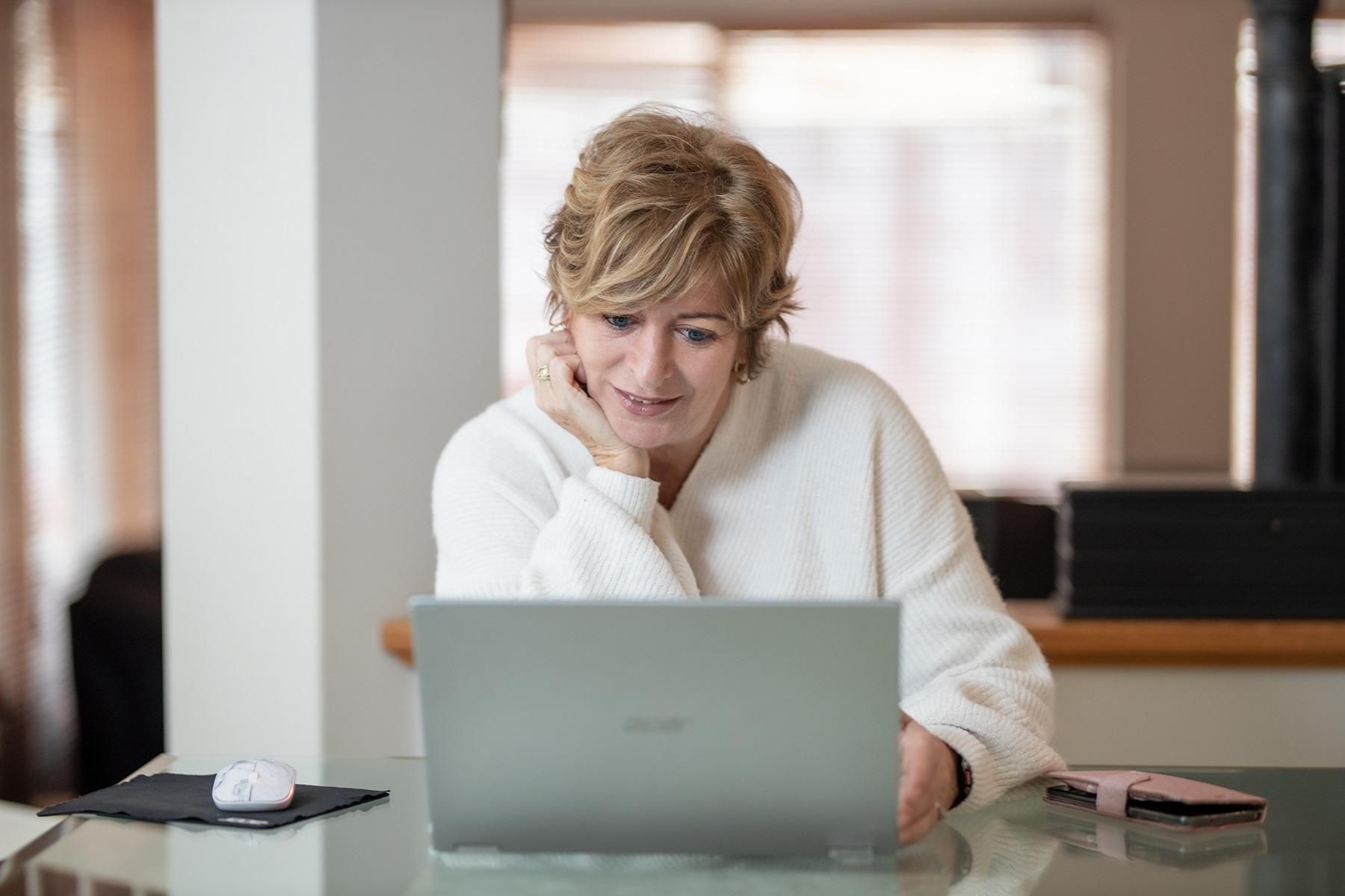 Photo of a woman working from her home-based business. She is sitting at a table and reading from her laptop.