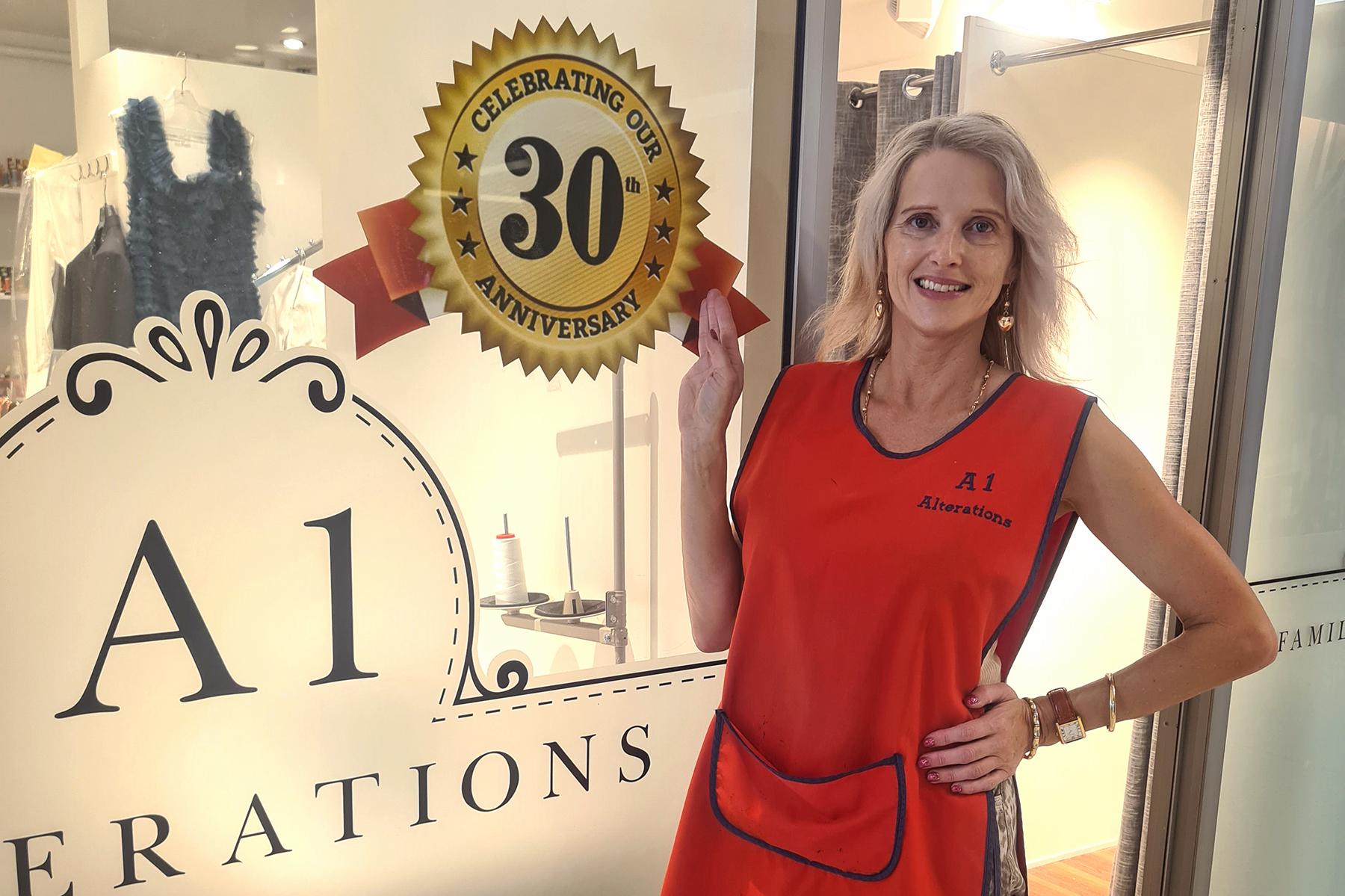 Photo of Fiona Larkin, owner of A1 Alterations, outside of her shop window. She is pointing to a sign that reads 'Celebrating our 30th anniversary'.