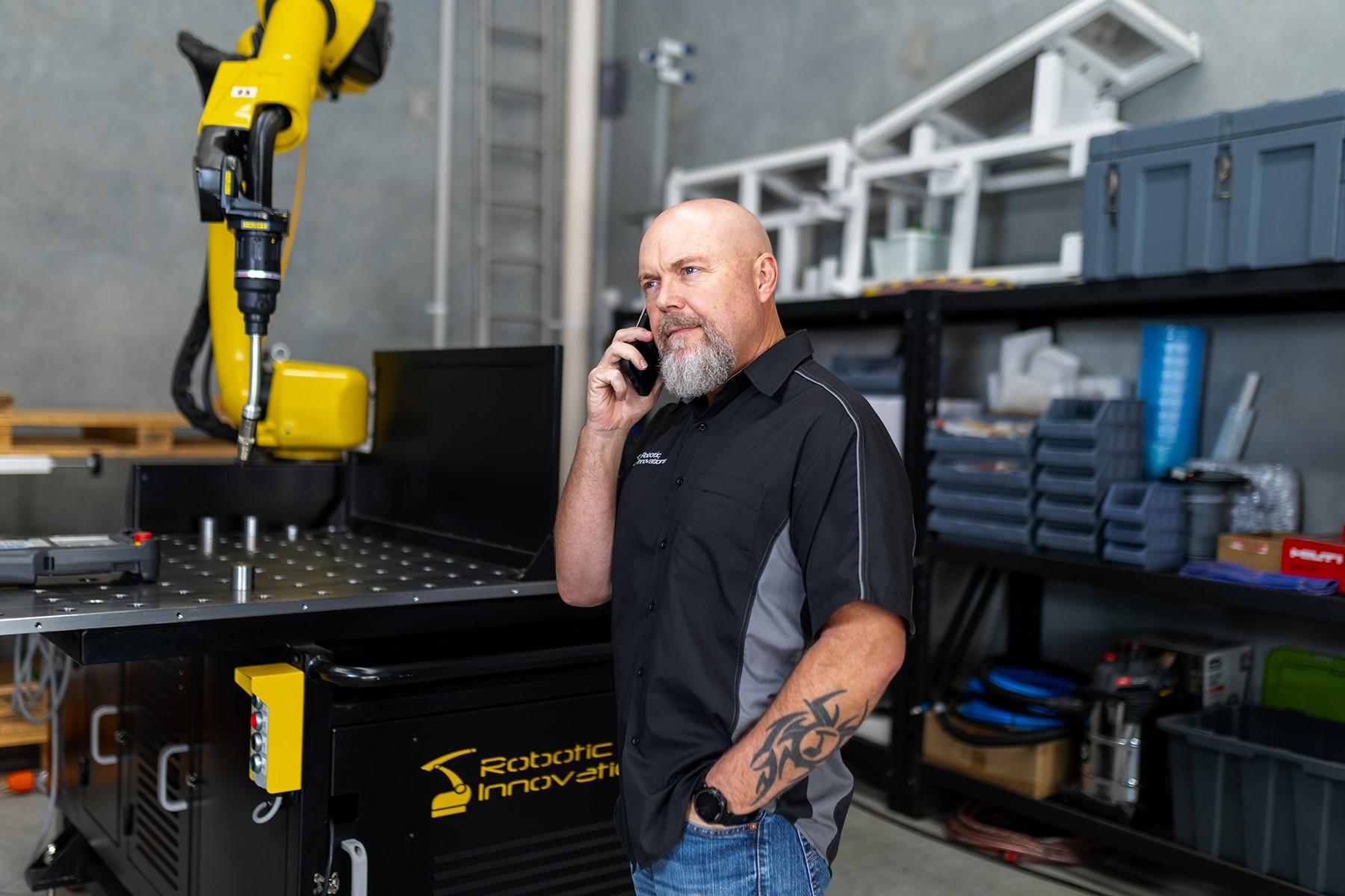 Photo of a man standing in a robotics warehouse. He is talking on a smartphone.
