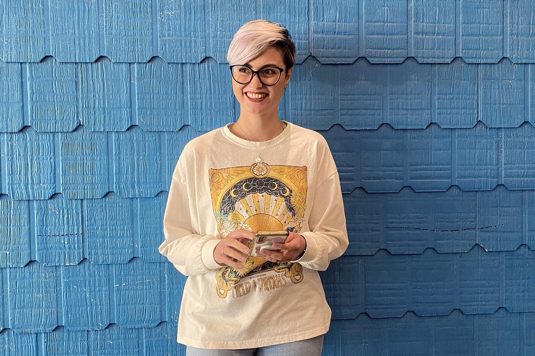 Photo of a woman standing against a wall. She is holding a smart phone and smiling.