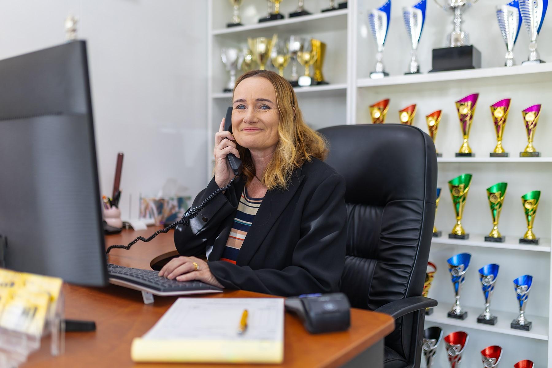 Photo of a woman in a trophy shop. She is sitting at a desk and holding a telephone to her ear.
