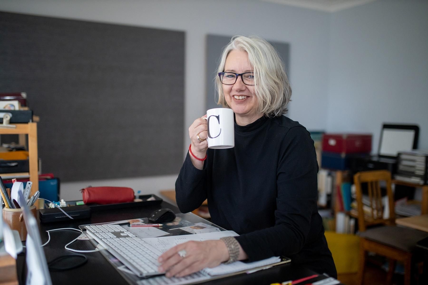 Photo of the owner of Fraulein Tech, Claudia Franke. She is sitting at a desk with a computer in front of her and is holding a cup of coffee. She is smiling at the camera.