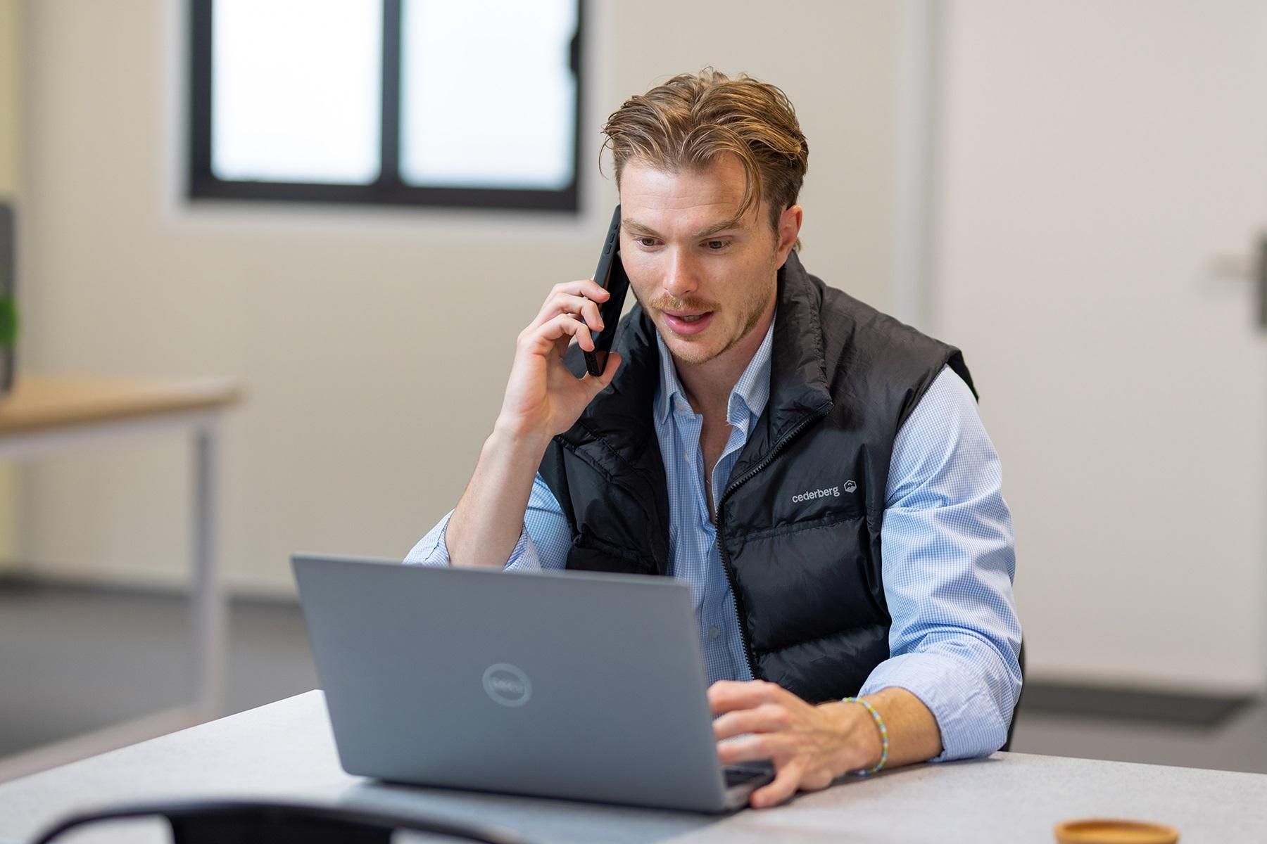 Photo of a male business owner sitting at a table. He is speaking on the phone and looking at his laptop.