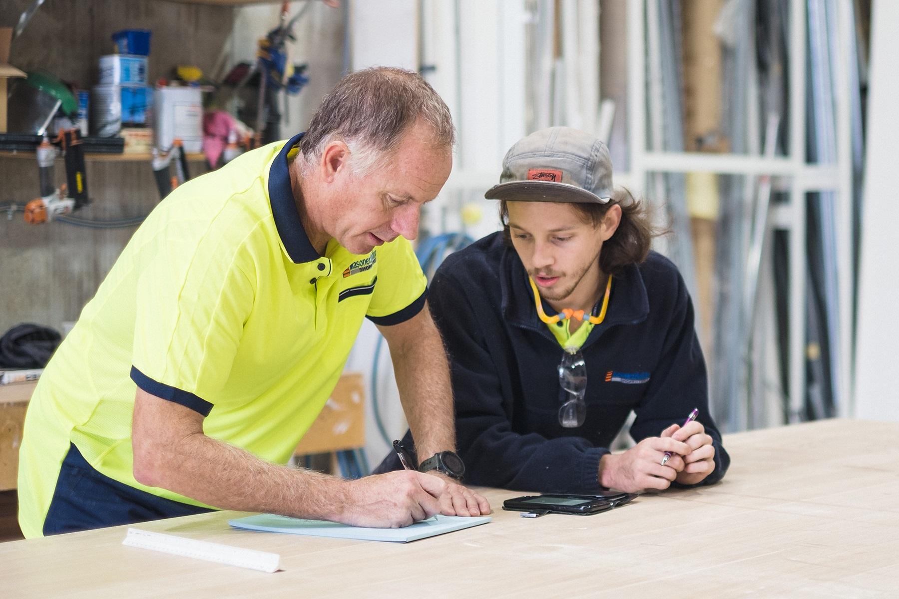A male business owner is writing in notebook as an employee looks on.