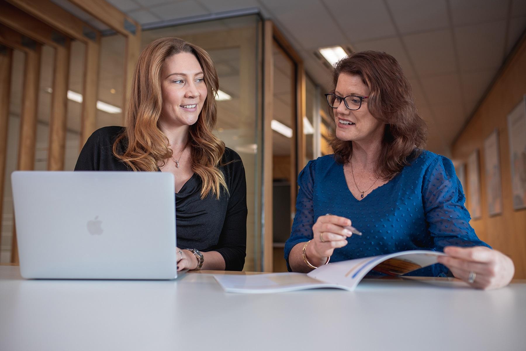 Photo of two women having a conversation. One is typing on a laptop and the other is holding documents.