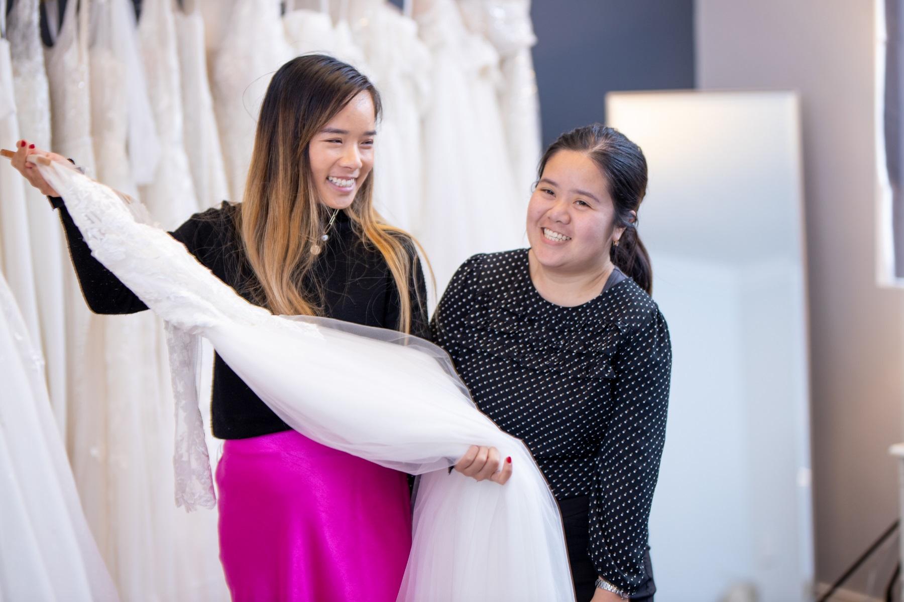 Photo of two female business owners in their bridal dress business. One is holding up a dress and both are smiling.