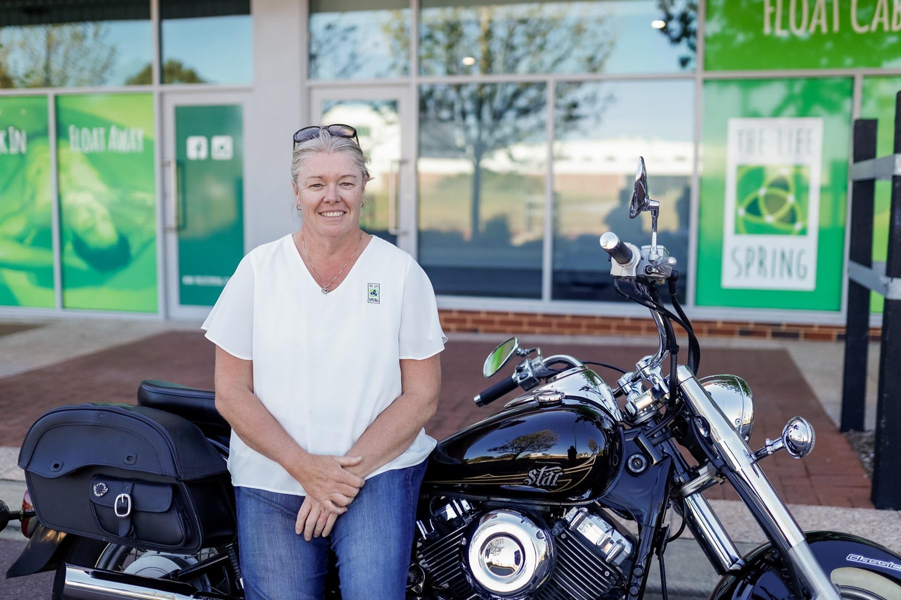Photo of a female business owner sitting on a motorcycle which is parked out the front of her business premises.