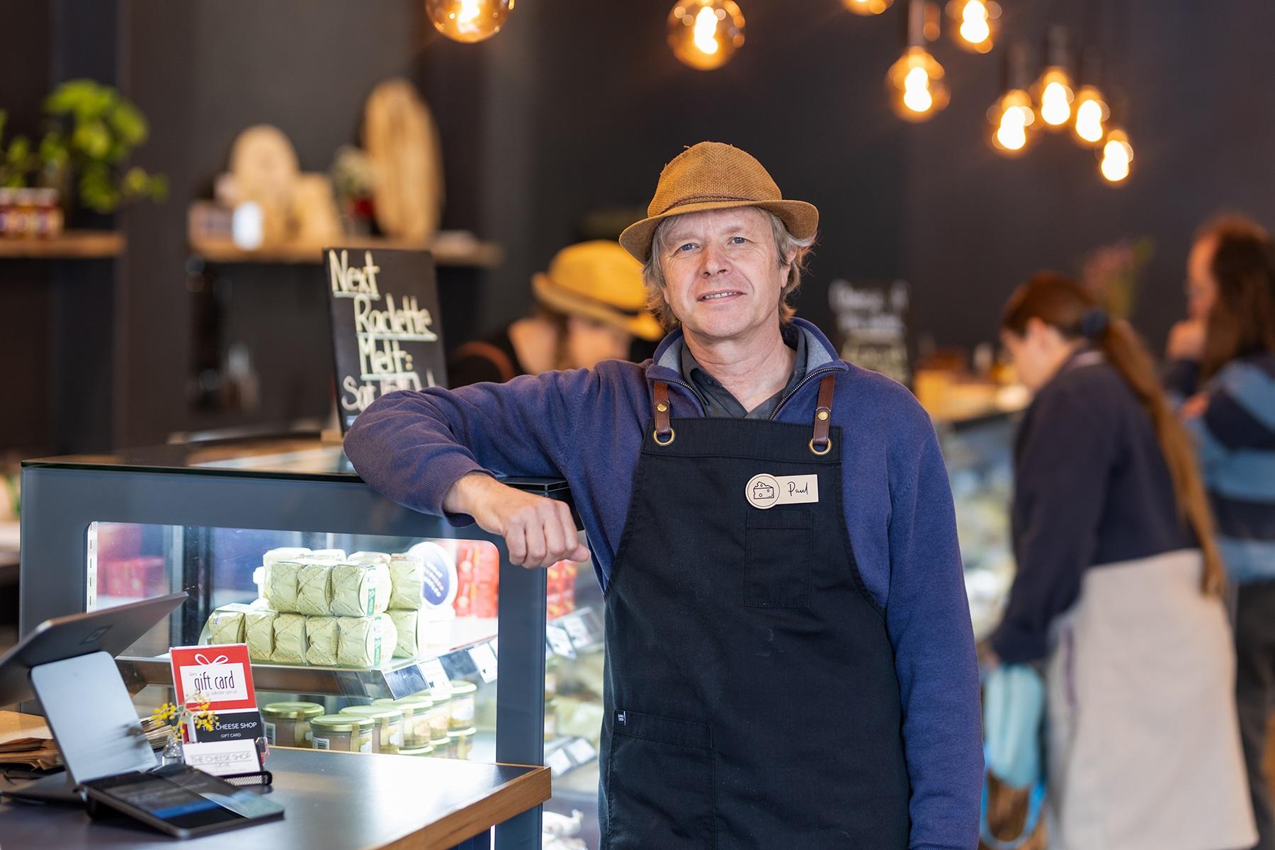 Photo of a male business owner. He is standing in his food business and has his arm up, leaning on a display fridge.