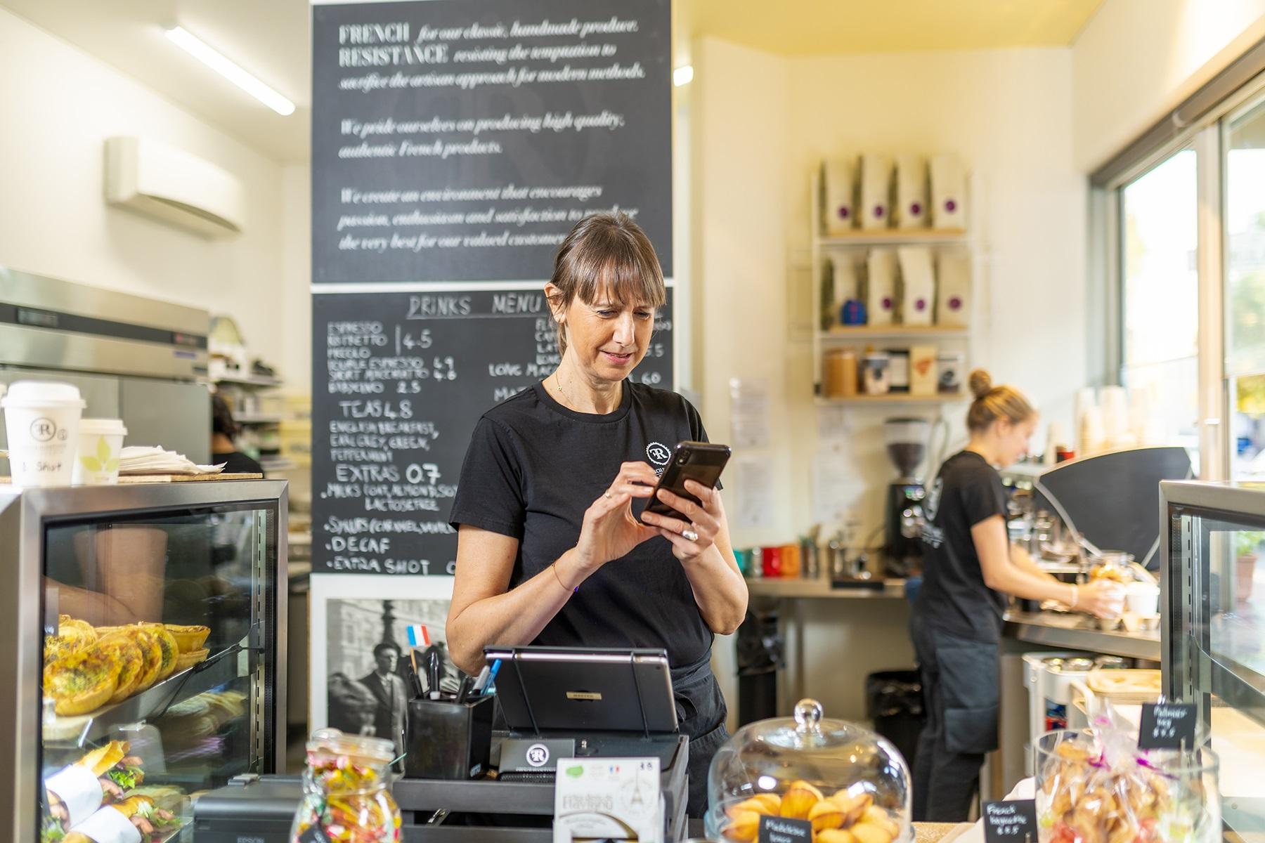 Photo of a female business owner in her food business. She is standing behind the shop counter and reading from her mobile phone.