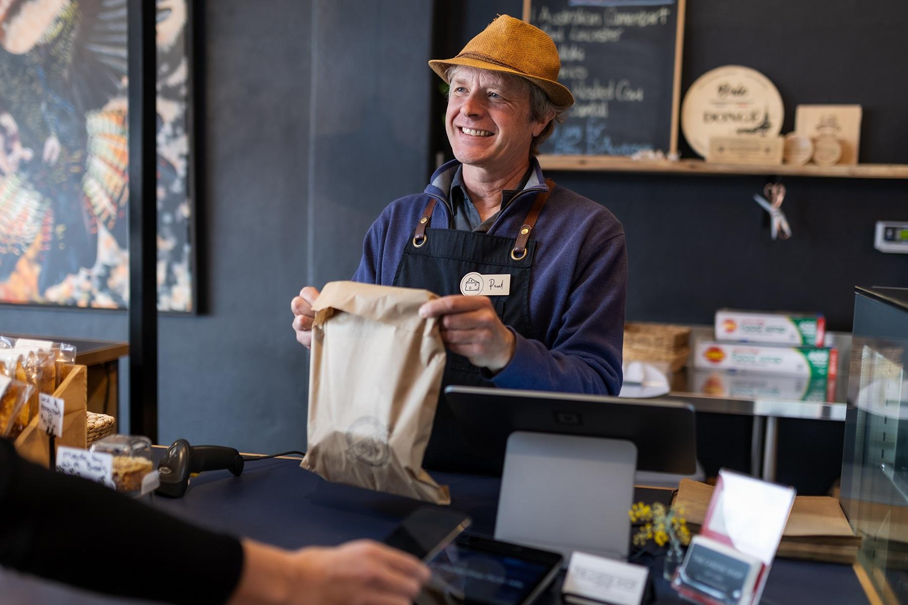 Photo of a male business owner. He is standing behind the counter of his retail store, serving a customer.