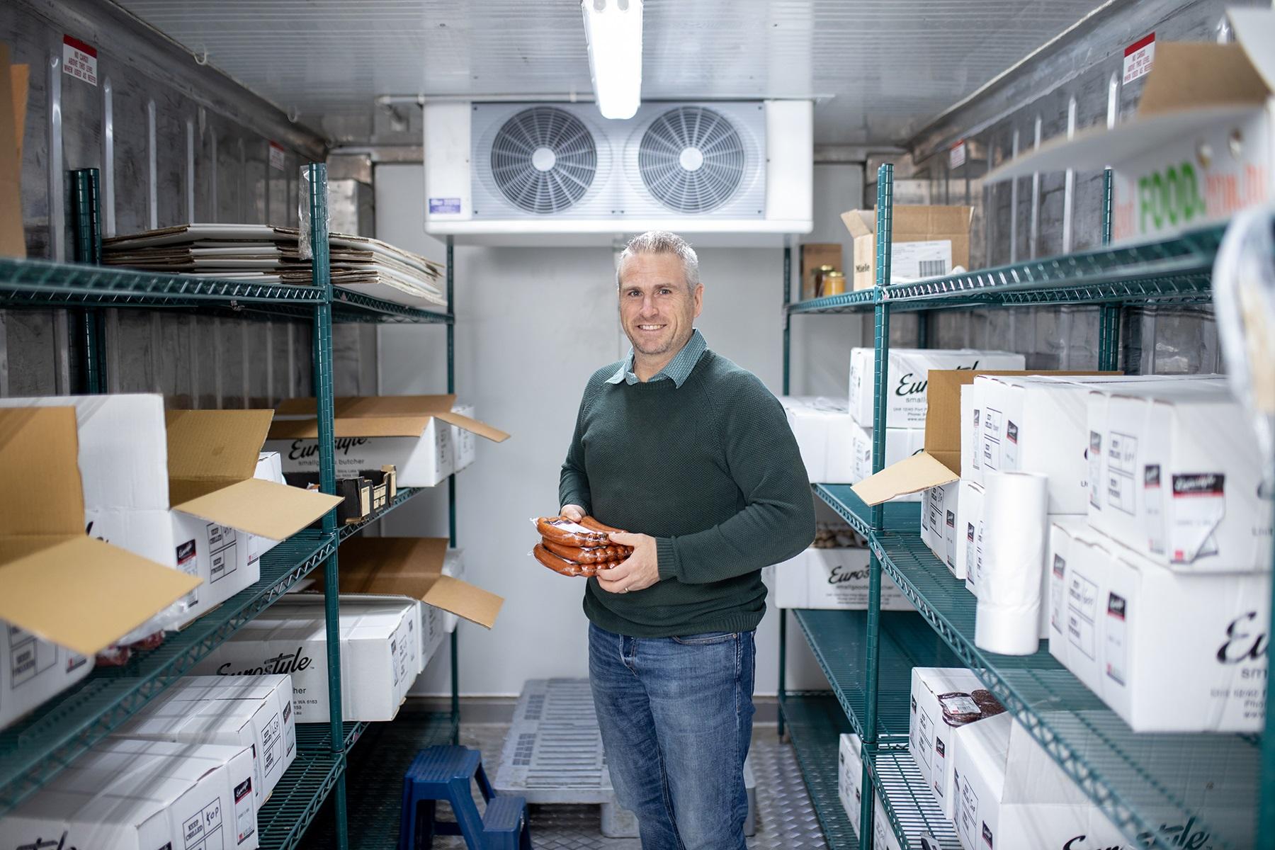 Photo of a man standing in a walk in freezer. He is holding packets of sausages in his hands.