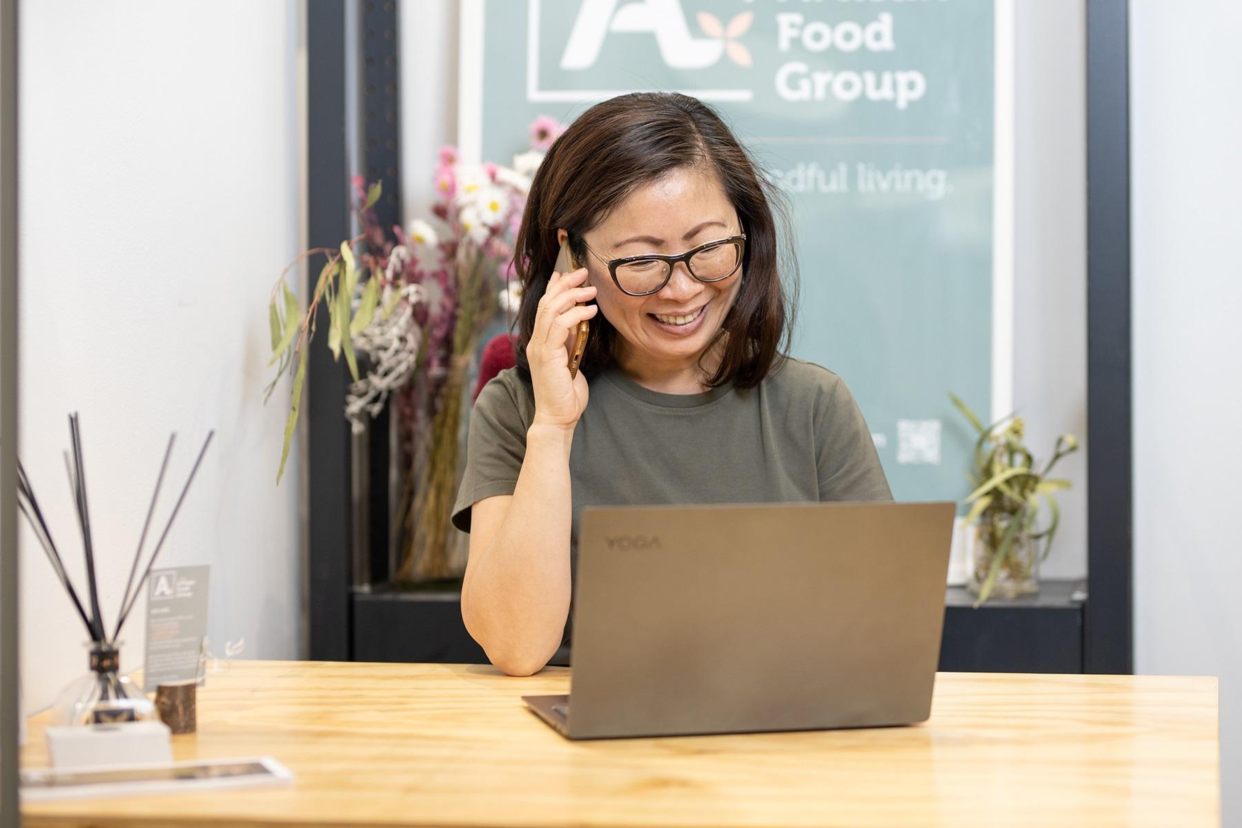 Photo of a woman sitting at a table and using a laptop while she speaks on the phone.
